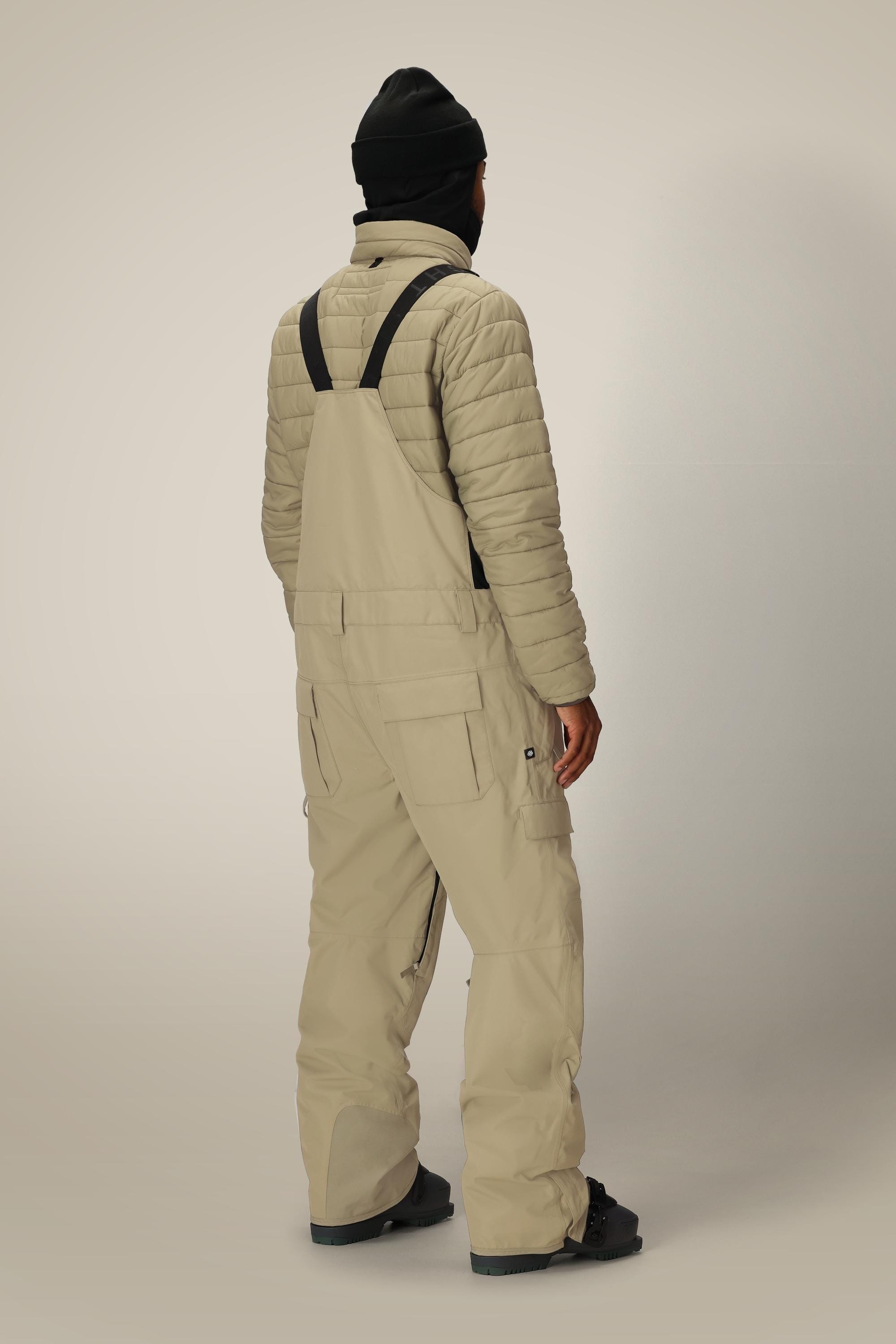 A person stands facing away, showcasing the back of their winter look in a black beanie, light quilted jacket, and 686 Mens 20K Hot Lap Shell Bib by 686 against a plain background.