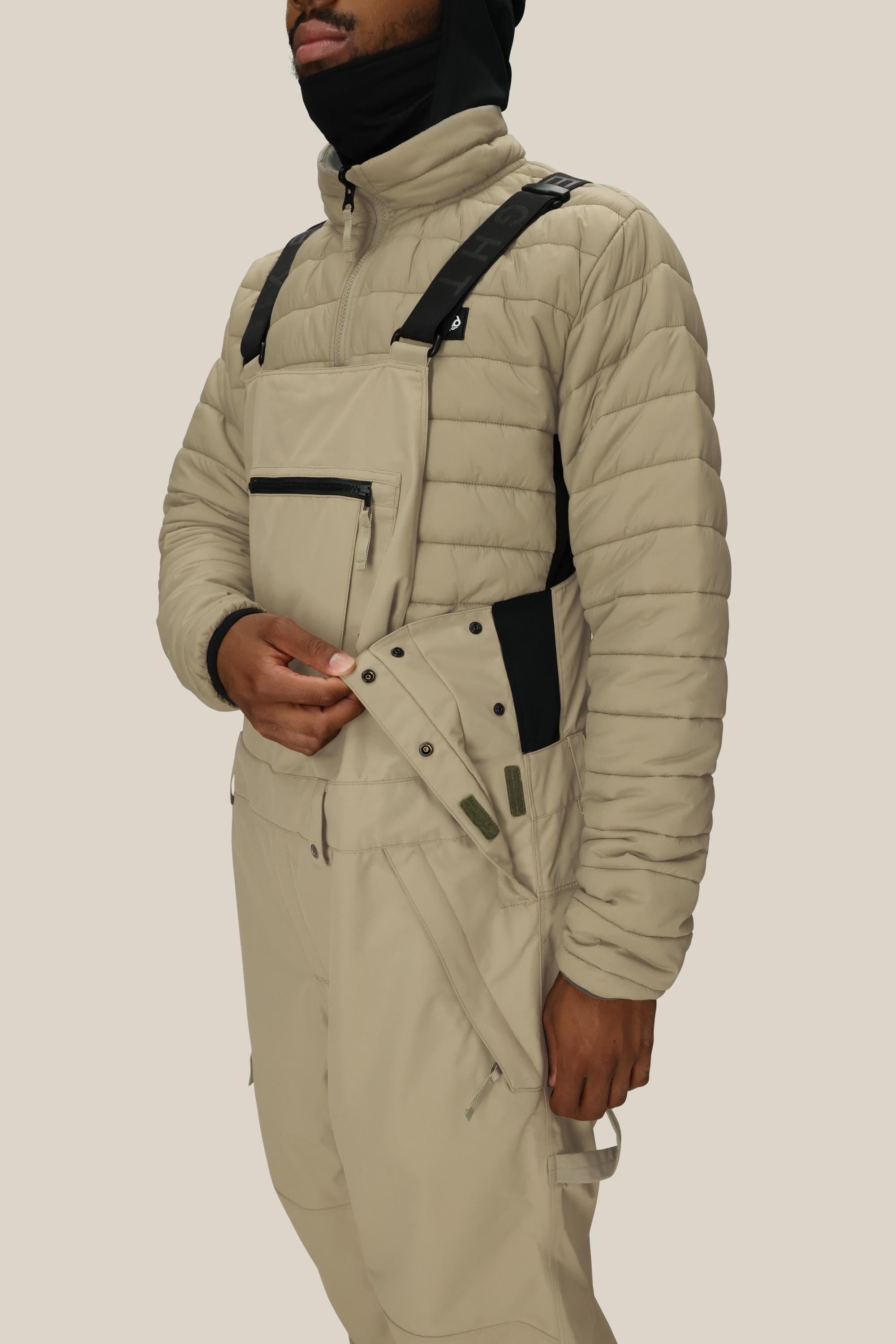 A person adjusts the side snaps of the 686 Mens 20K Hot Lap Shell Bib by 686, wearing a beige quilted jacket and matching bib overalls with black straps. The background is plain and neutral.