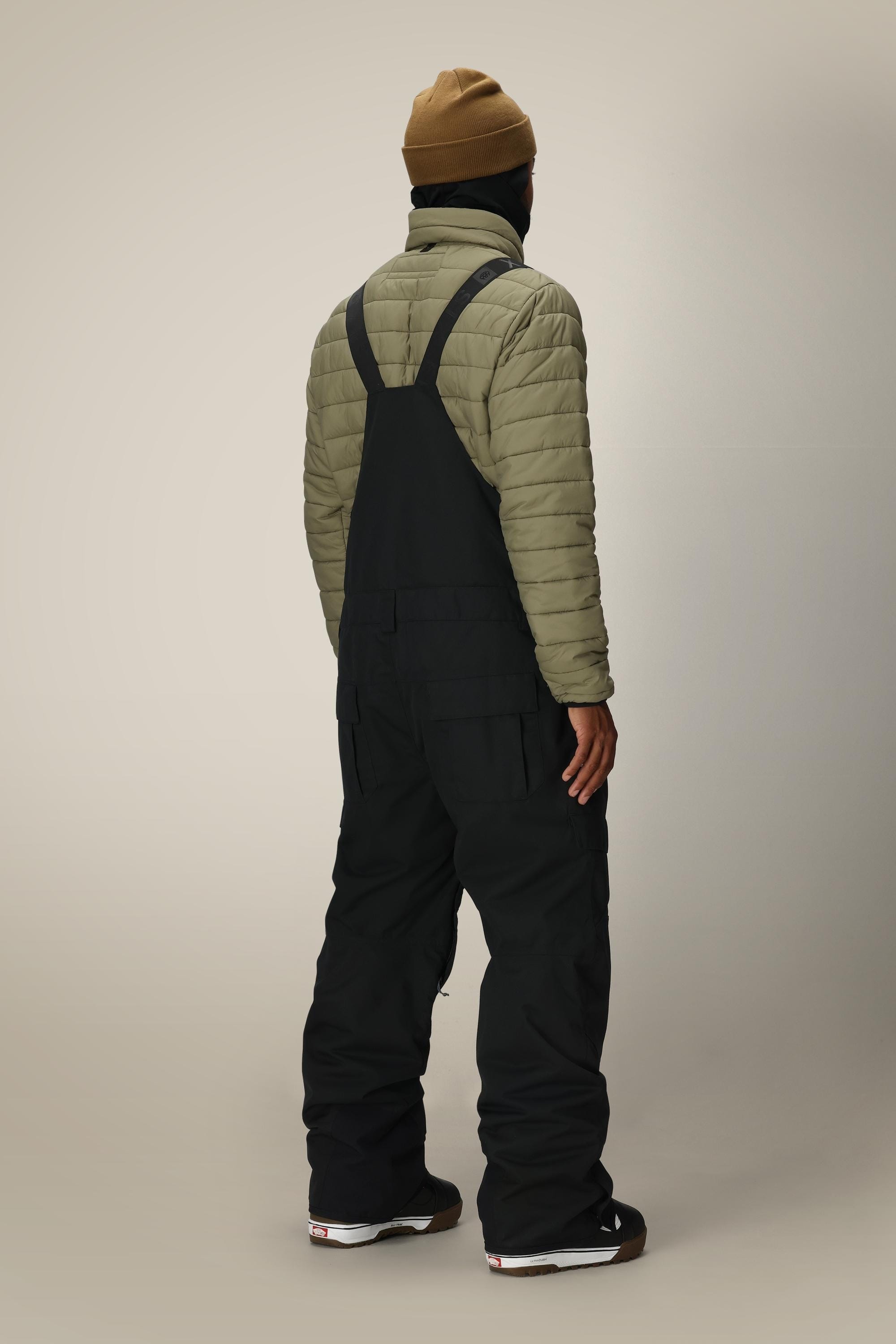 A person stands facing away, wearing a brown beanie, olive puffer jacket, and the 686 Men’s Hot Lap™ Insulated Bib in black with black shoes, against a plain light background.