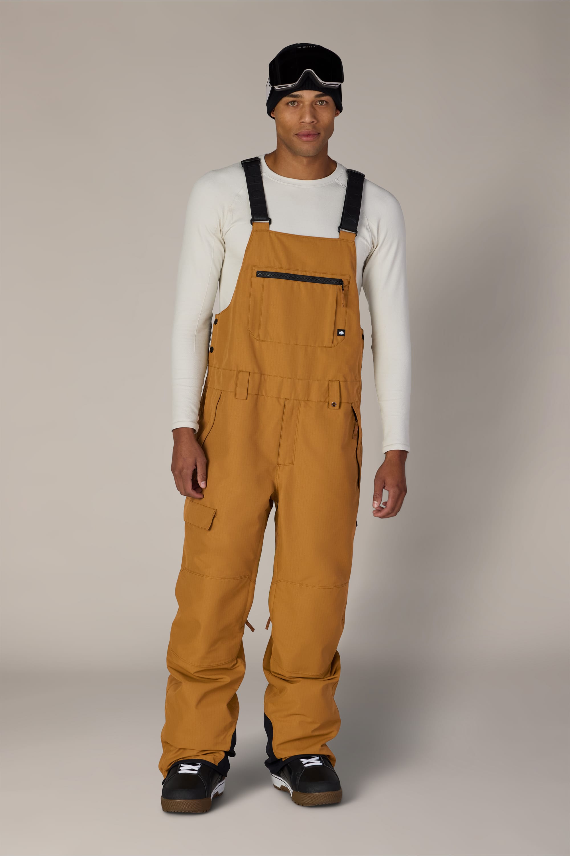 A man stands facing forward, wearing the 686 Men’s Hot Lap™ Insulated Bib by 686 in tan, paired with a white long-sleeve shirt, black snow boots, and a black ski mask pushed up on his forehead. The background is plain and neutral.