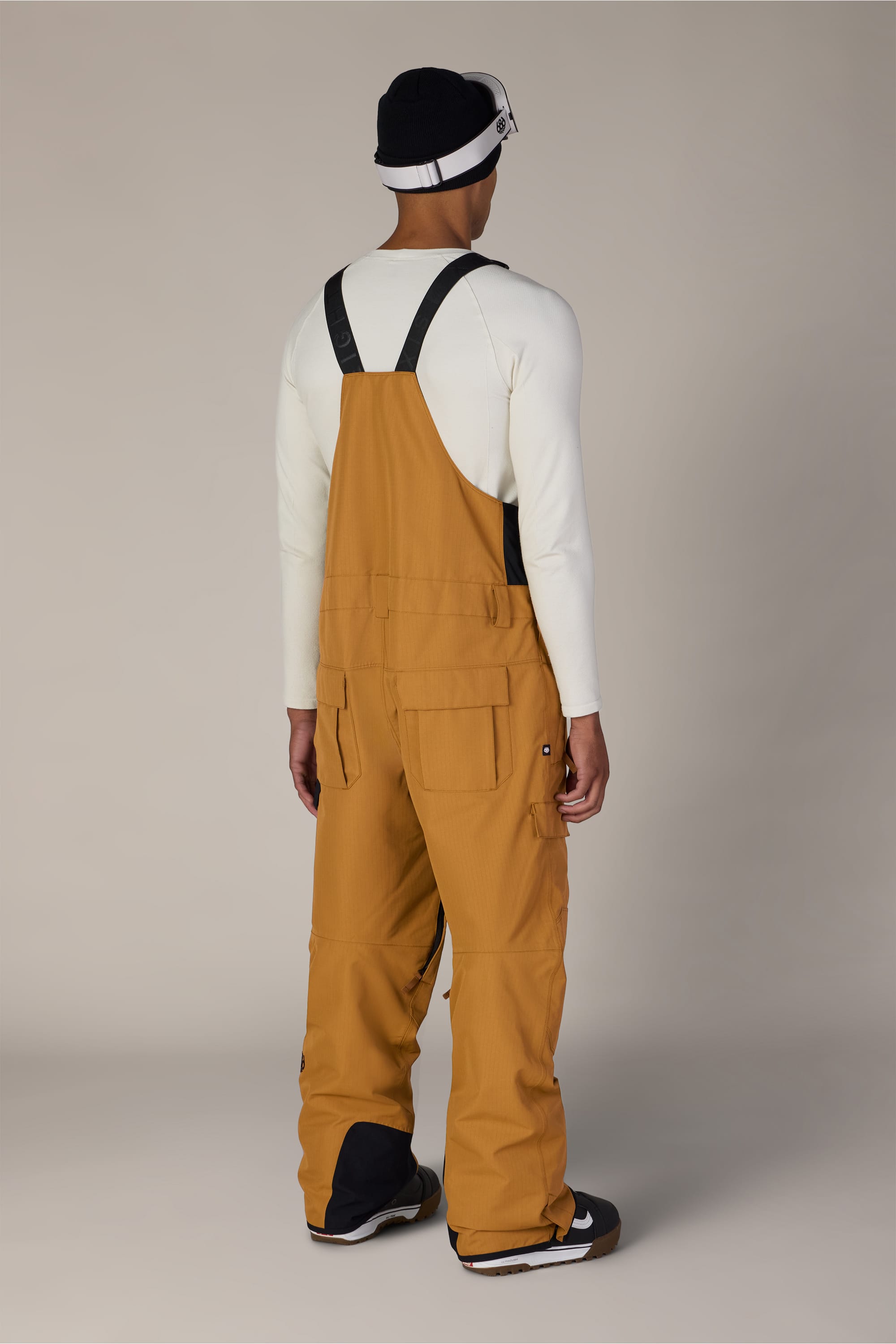 A person stands facing away against a plain light background, wearing the 686 Men’s Hot Lap™ Insulated Bib in mustard yellow, paired with a white long-sleeve shirt, a black-and-white beanie, and black sneakers with white soles.