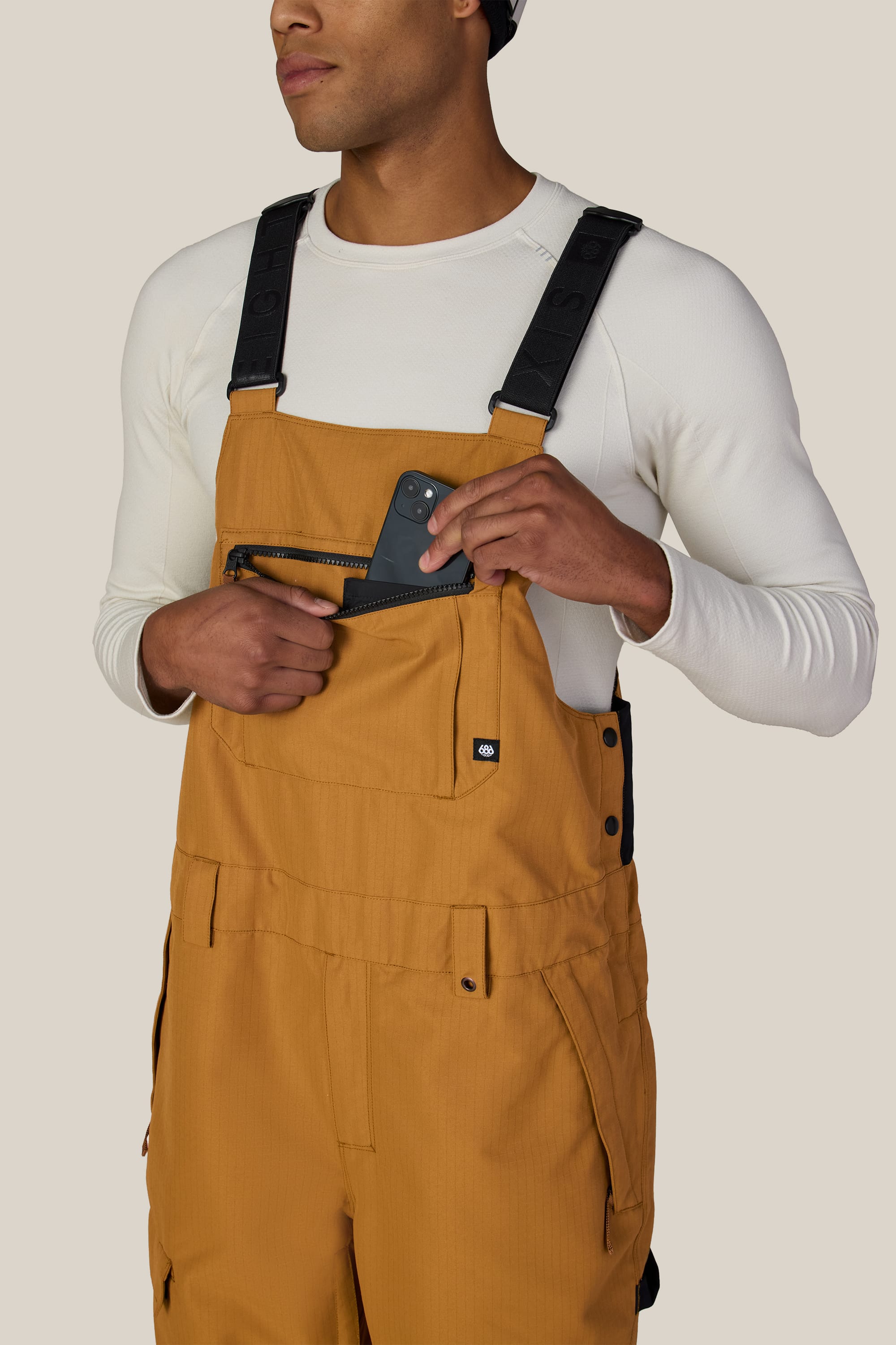 A person wearing the 686 Men’s Hot Lap™ Insulated Bib in tan and a white long-sleeve shirt places a black smartphone into the zippered chest pocket. The background is plain beige.