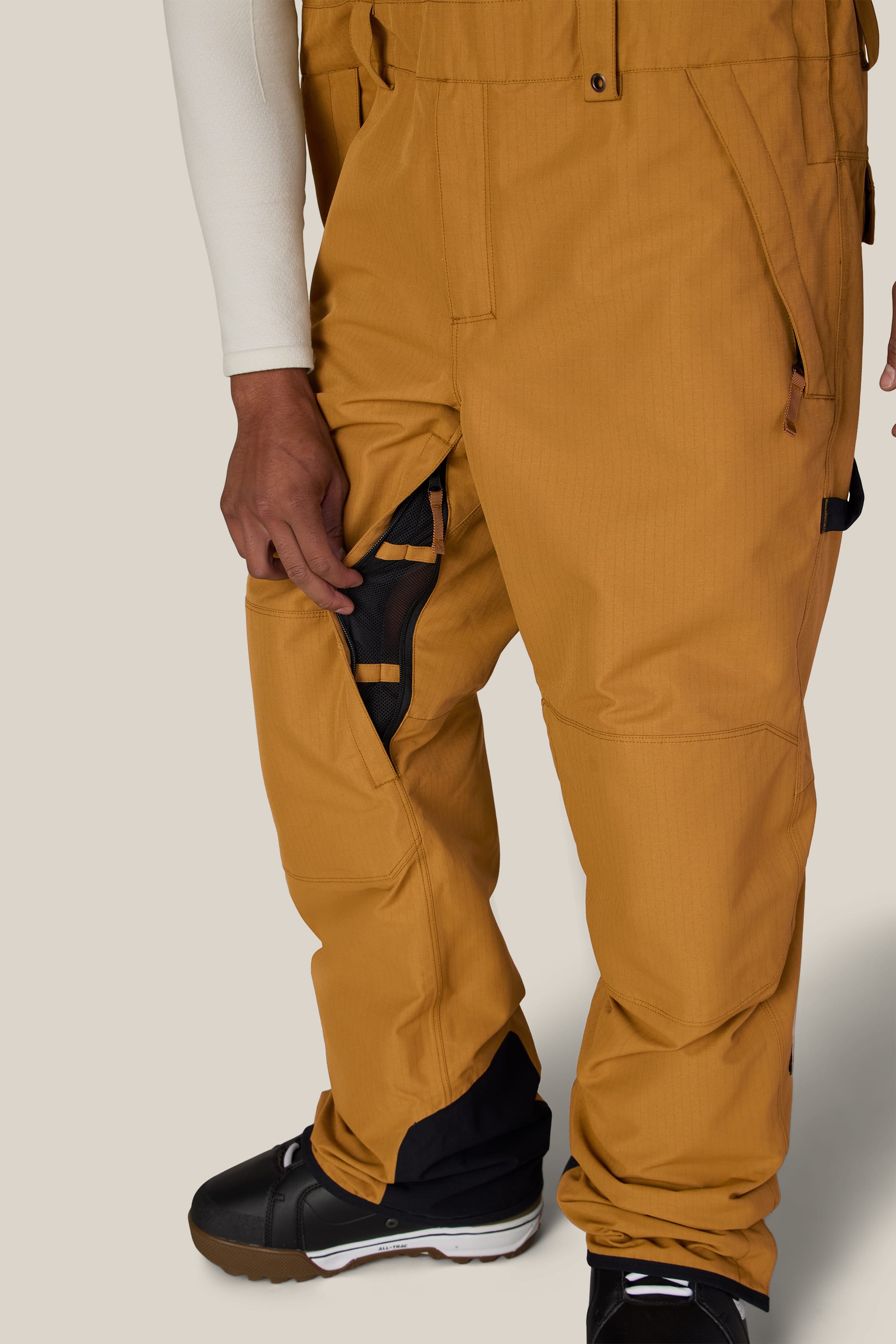 A person wearing the 686 Men’s Hot Lap™ Insulated Bib in tan, paired with a white long-sleeve shirt and black snow boots, stands on a beige background. Their hand rests near the zippered side pocket of the 686 bib.