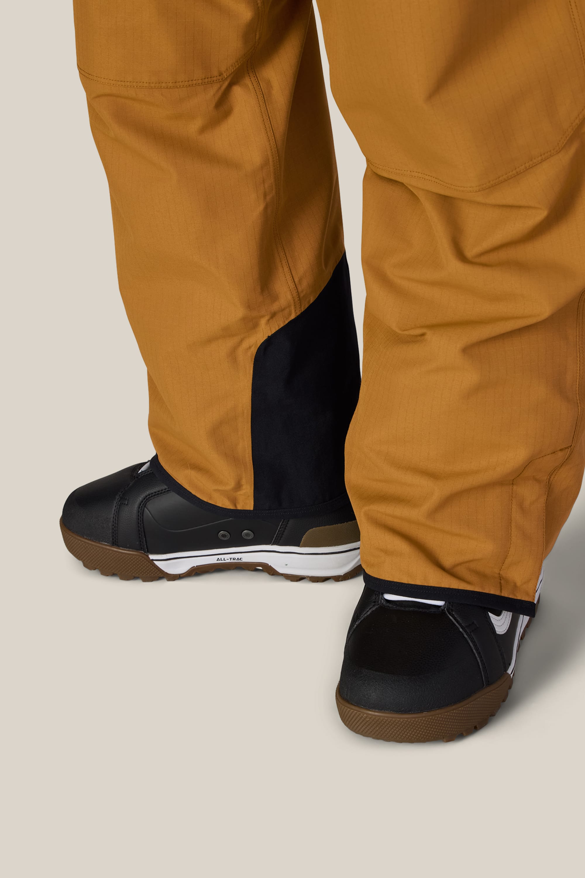 A close-up of a person wearing the 686 Men’s Hot Lap™ Insulated Bib by 686, featuring brown fabric with black patches over the heels, paired with black snow boots, standing on a beige background.