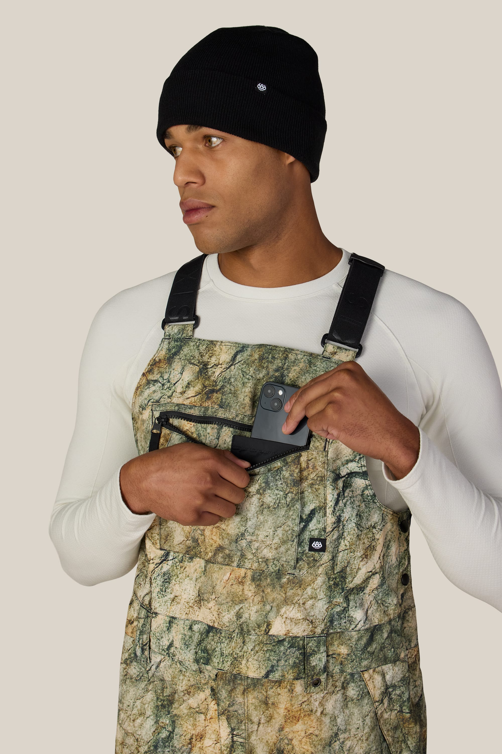 A man wearing a black beanie and white long-sleeve shirt places a black smartphone into the front pocket of his green camo 686 Men’s Hot Lap™ Insulated Bib by 686 while looking to the side.