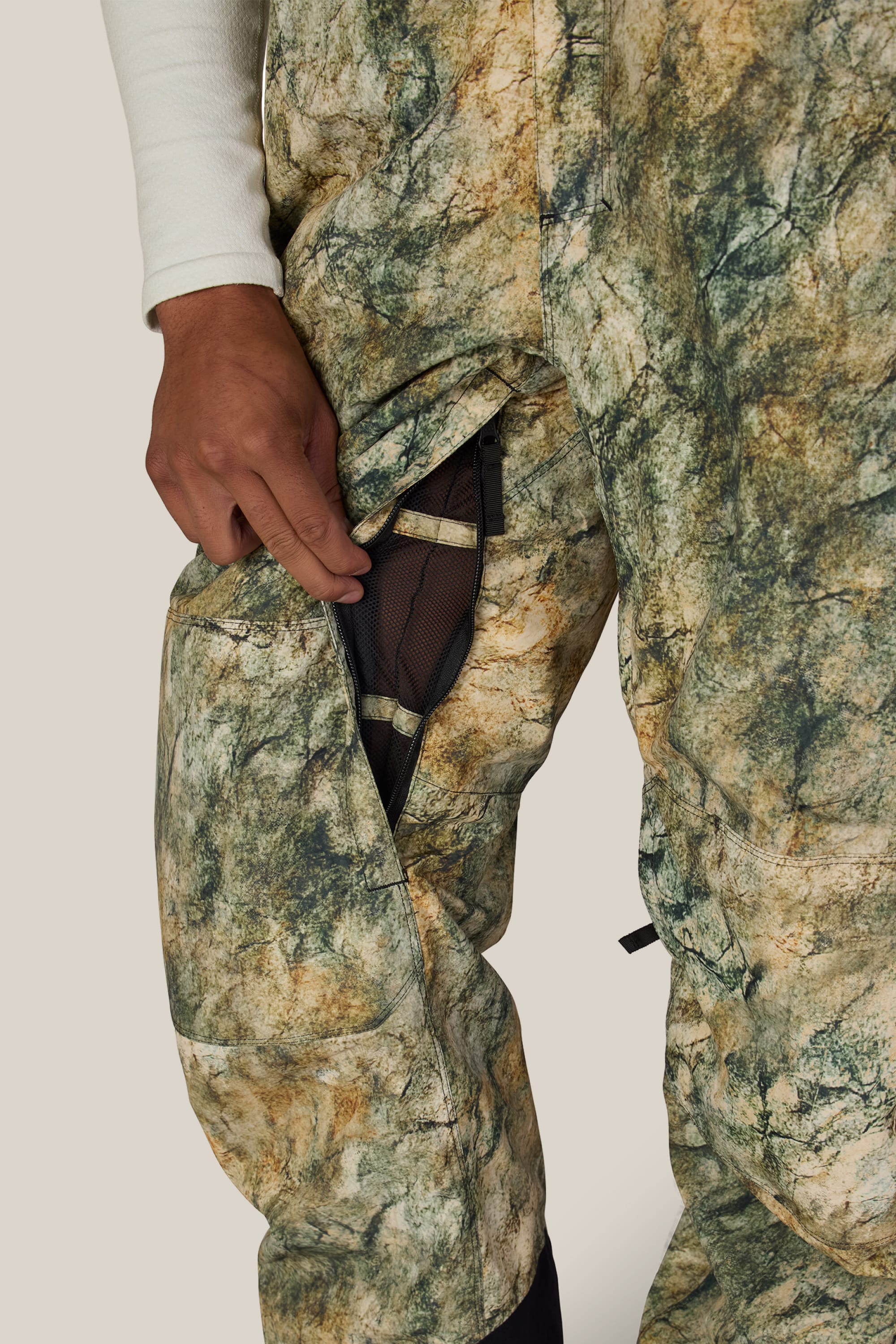 A person wearing the 686 Men’s Hot Lap™ Insulated Bib in camouflage opens a zippered mesh pocket on the side of their thigh against a plain, neutral background.