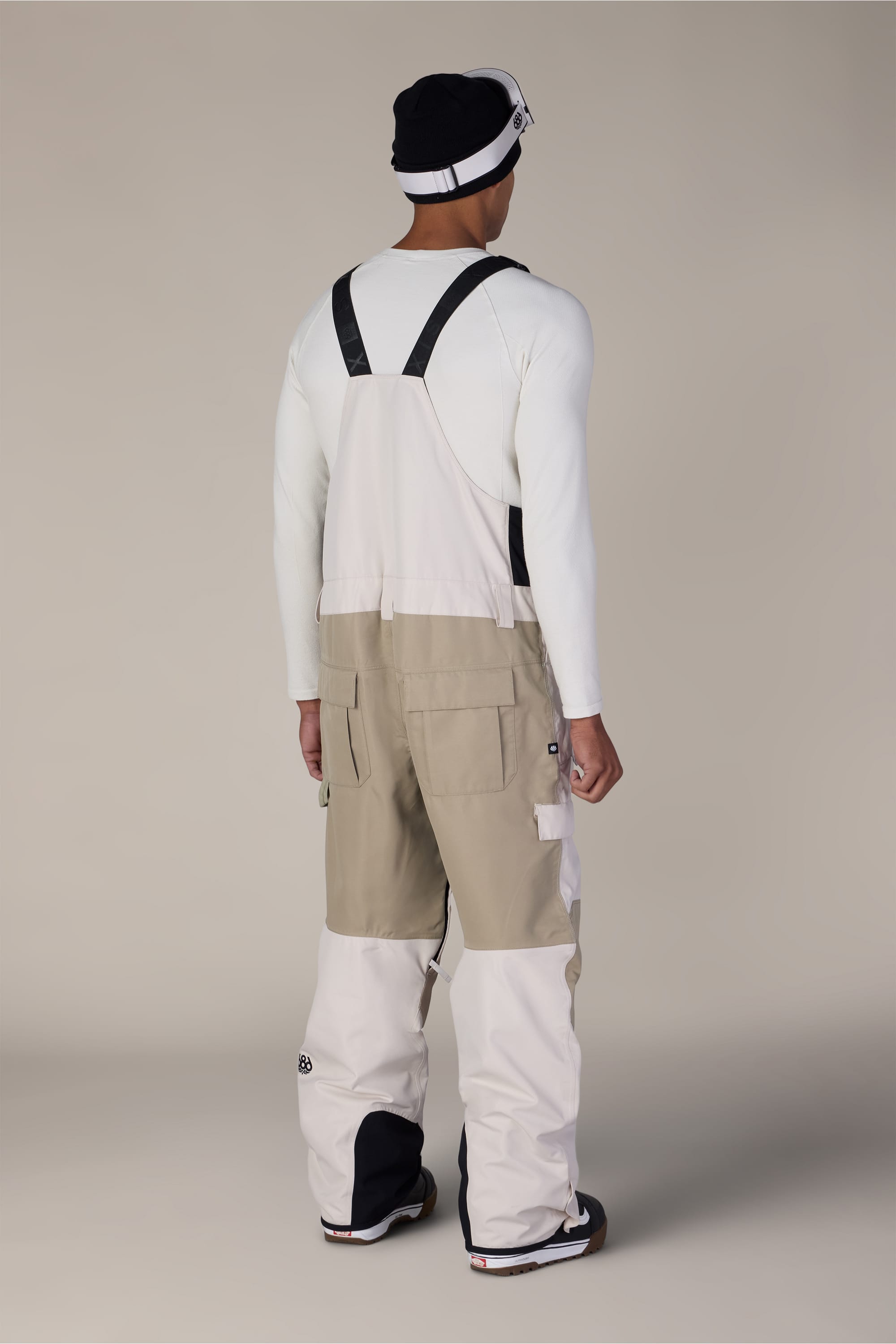 A person stands with their back to the camera against a plain background, wearing a white long-sleeve shirt, snow boots, a black and white beanie, and the 686 Men’s Hot Lap™ Insulated Bib by 686 in beige and white with black straps.