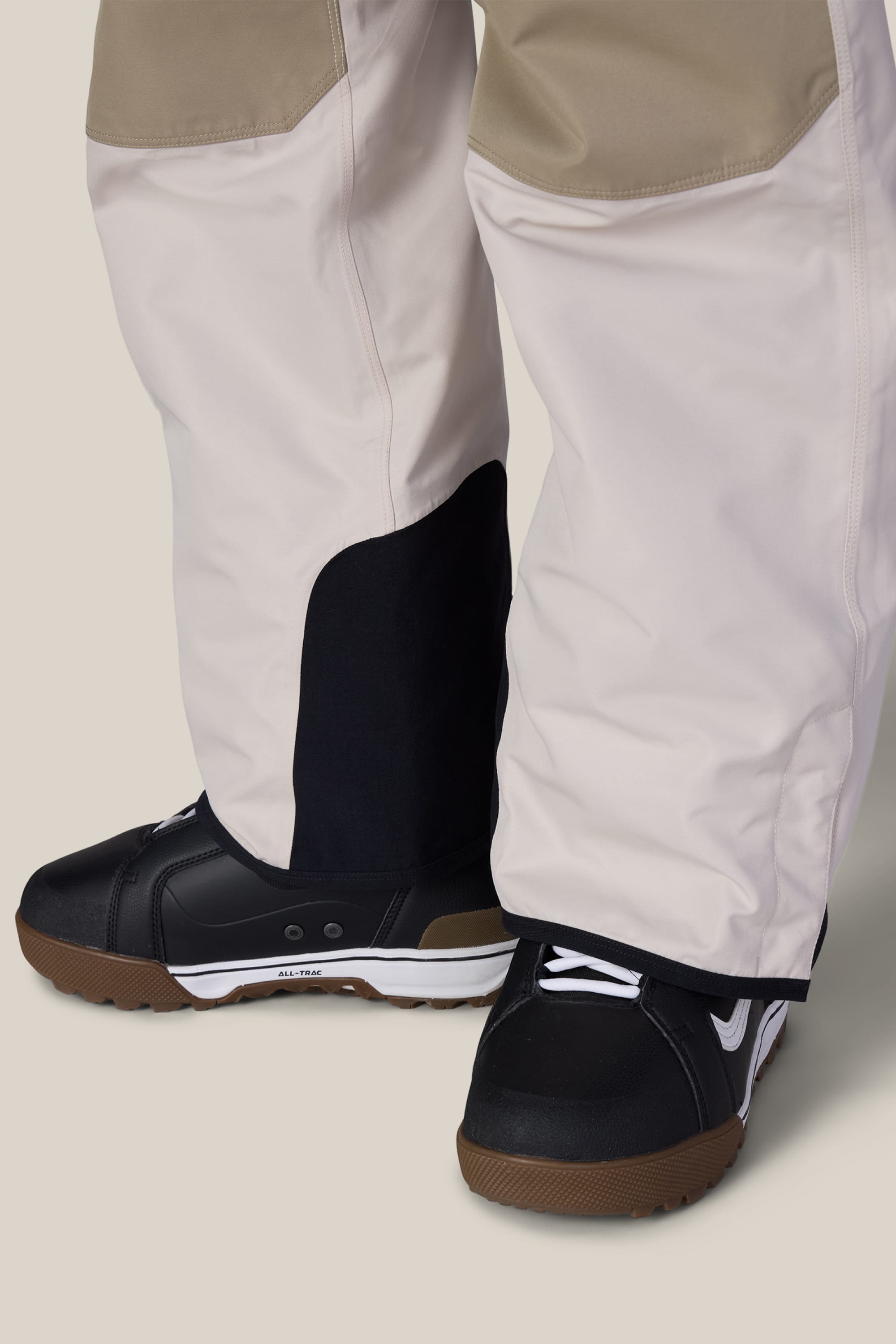 Close-up of a person wearing the 686 Men’s Hot Lap™ Insulated Bib in beige and white, featuring black patches near the ankles, paired with black sneakers with white accents and brown soles. The background is plain and light-colored.