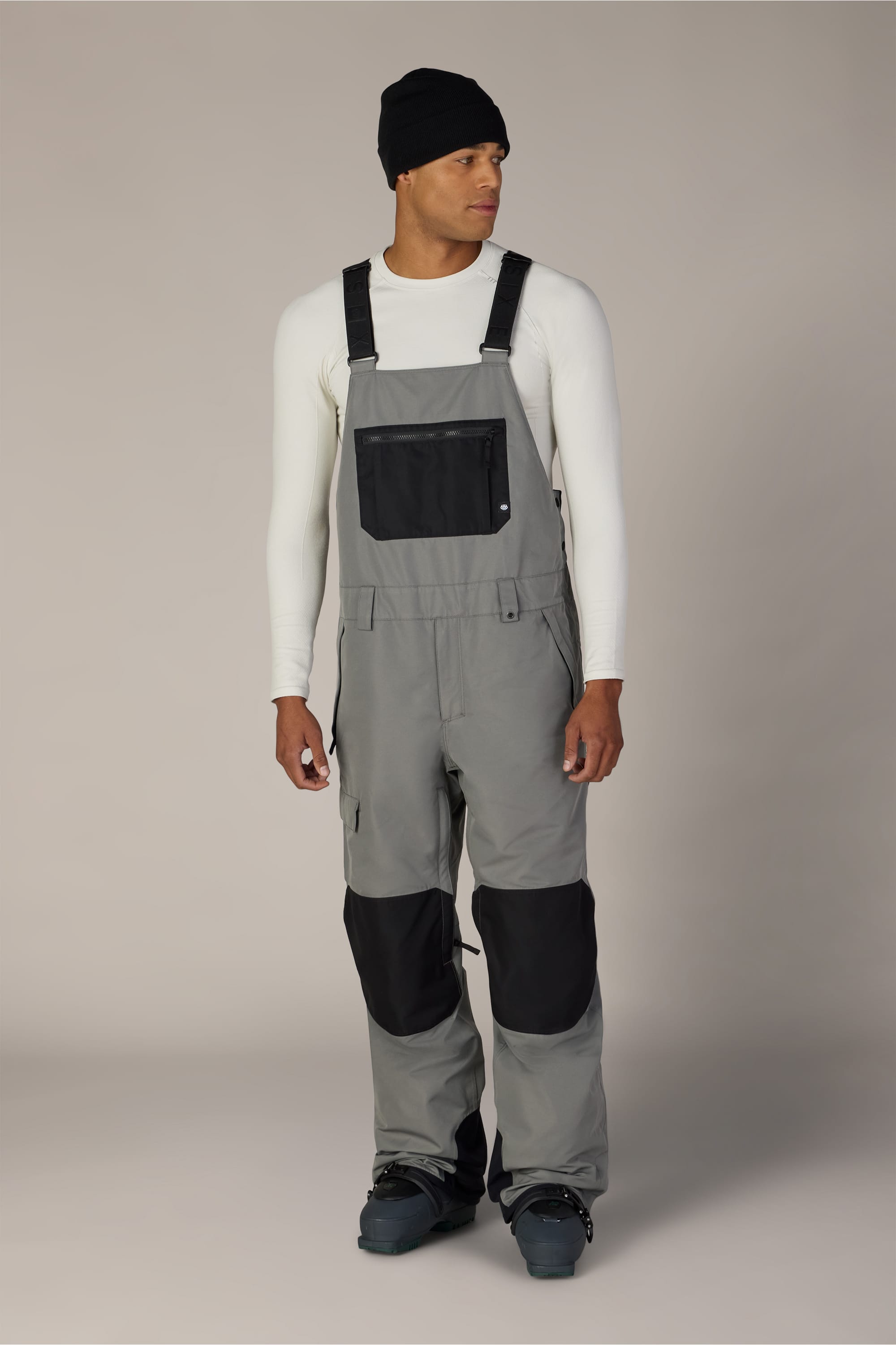 A man wearing the 686 Men’s Hot Lap™ Insulated Bib by 686—characterized by its gray color, black knee patches, and large chest pocket—paired with a black beanie, white long-sleeve shirt, and dark boots stands slightly turned to the right against a plain background.