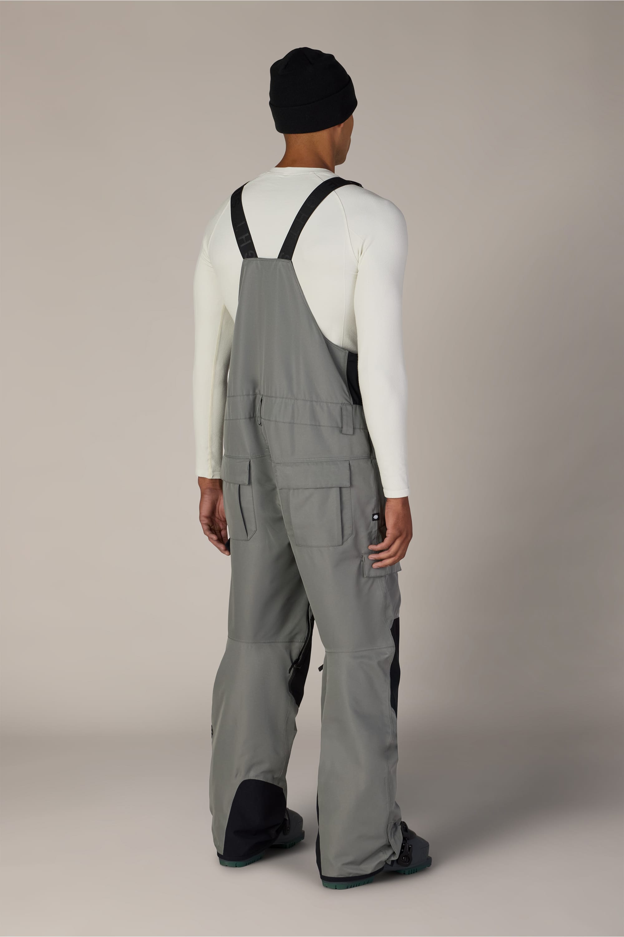 A person stands facing away, wearing a black beanie and a white long-sleeve shirt, paired with the 686 Men’s Hot Lap™ Insulated Bib by 686—a gray bib overall featuring black straps and black patches on the lower legs—set against a plain background.