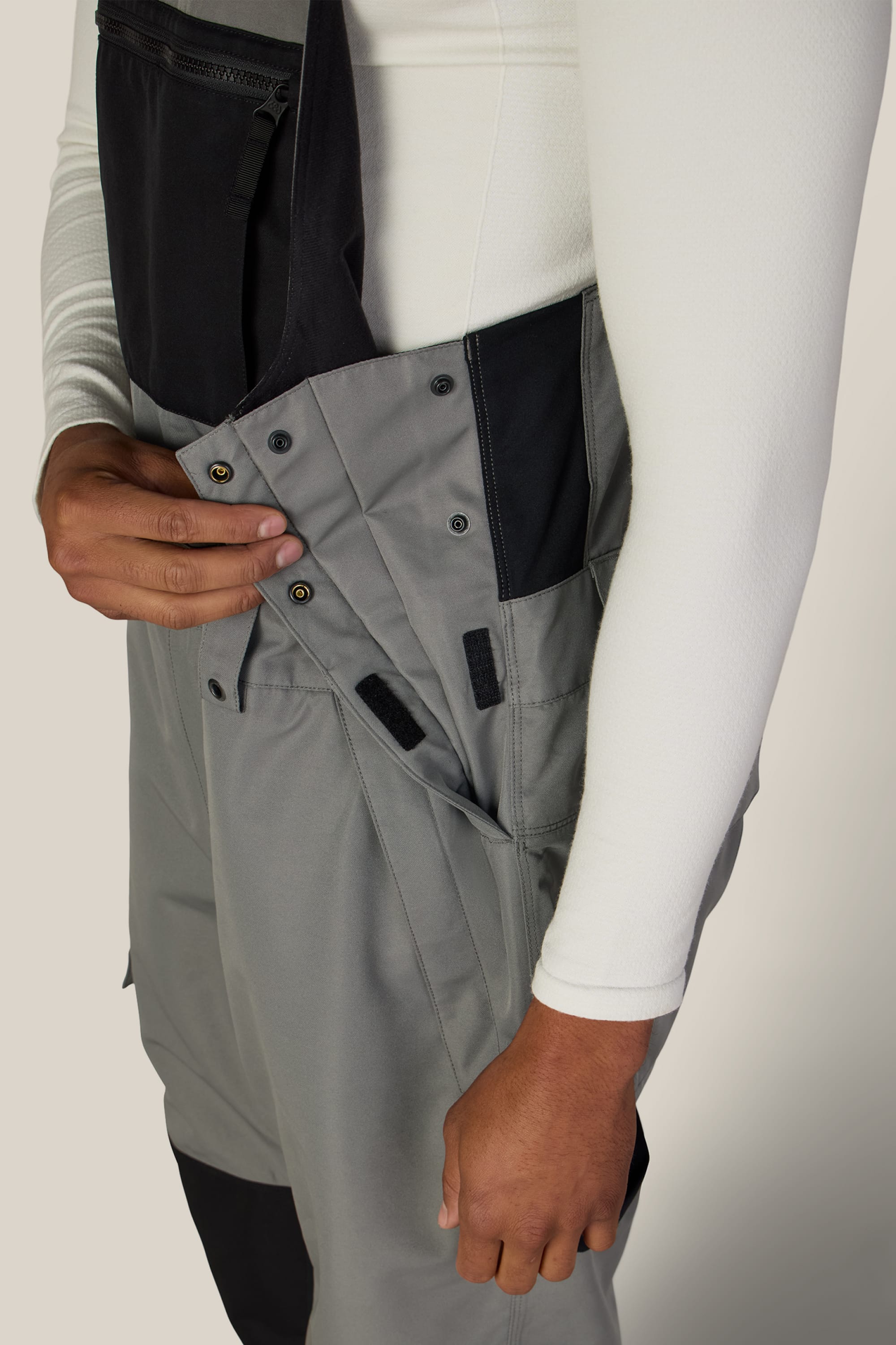 A person wearing a white long-sleeve shirt fastens the side buttons of the 686 Men’s Hot Lap™ Insulated Bib by 686, featuring gray fabric with black accents.