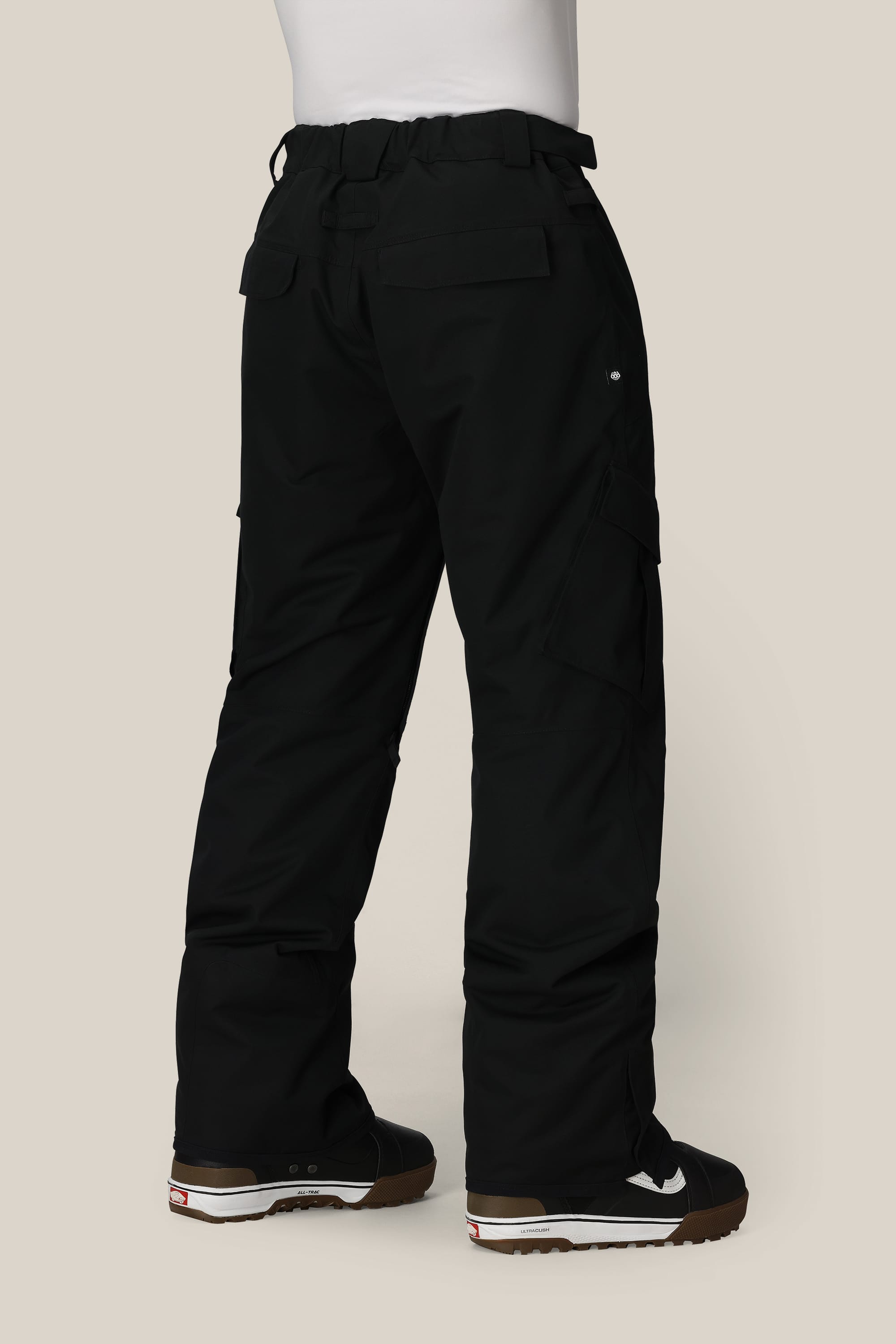 A person is seen from the back, wearing 686 Mens Infinity Insulated Cargo Pants in black and black sneakers with white soles, standing against a light beige background. The image focuses on the lower half of their body.