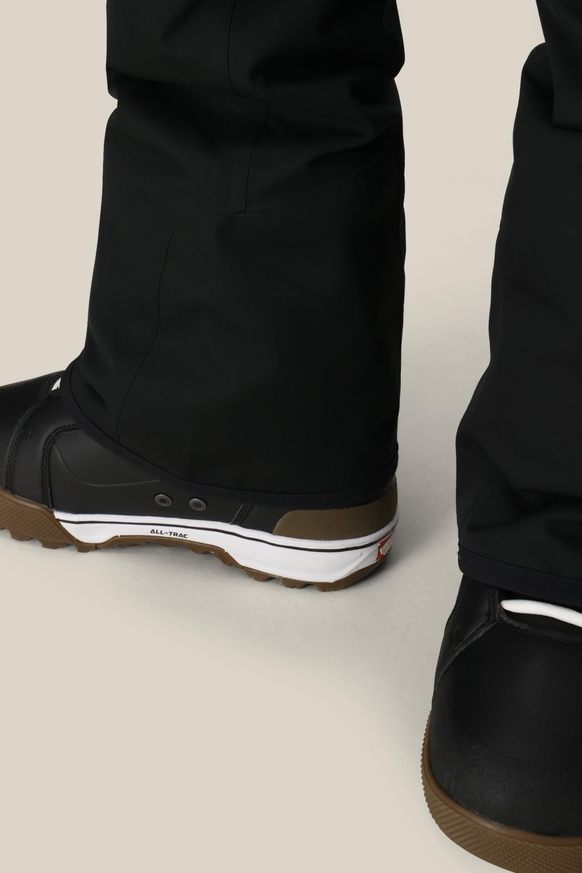 Close-up of a person wearing 686 Mens Infinity Insulated Cargo Pant and black snow boots with brown soles, standing on a light-colored surface. The pants from 686 partially cover the boots.
