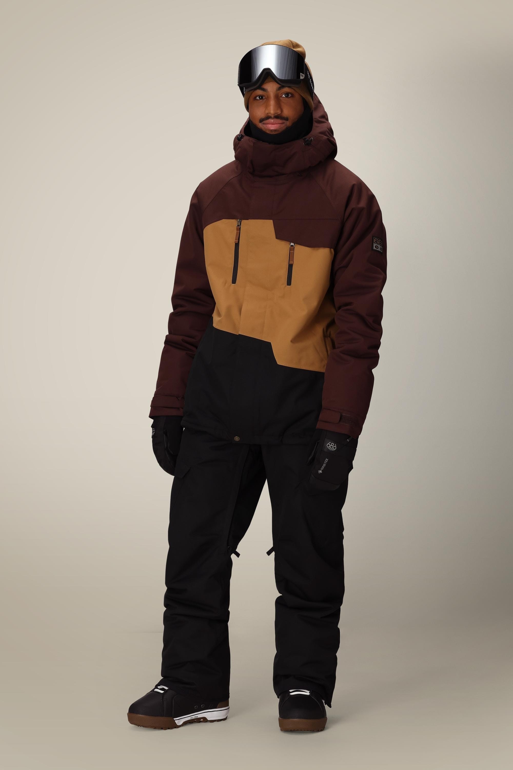 A person wearing 686 Mens Infinity Insulated Cargo Pant, a brown and tan winter jacket, black gloves, boots, and ski goggles stands against a plain background, ready for cold weather or snow activities.