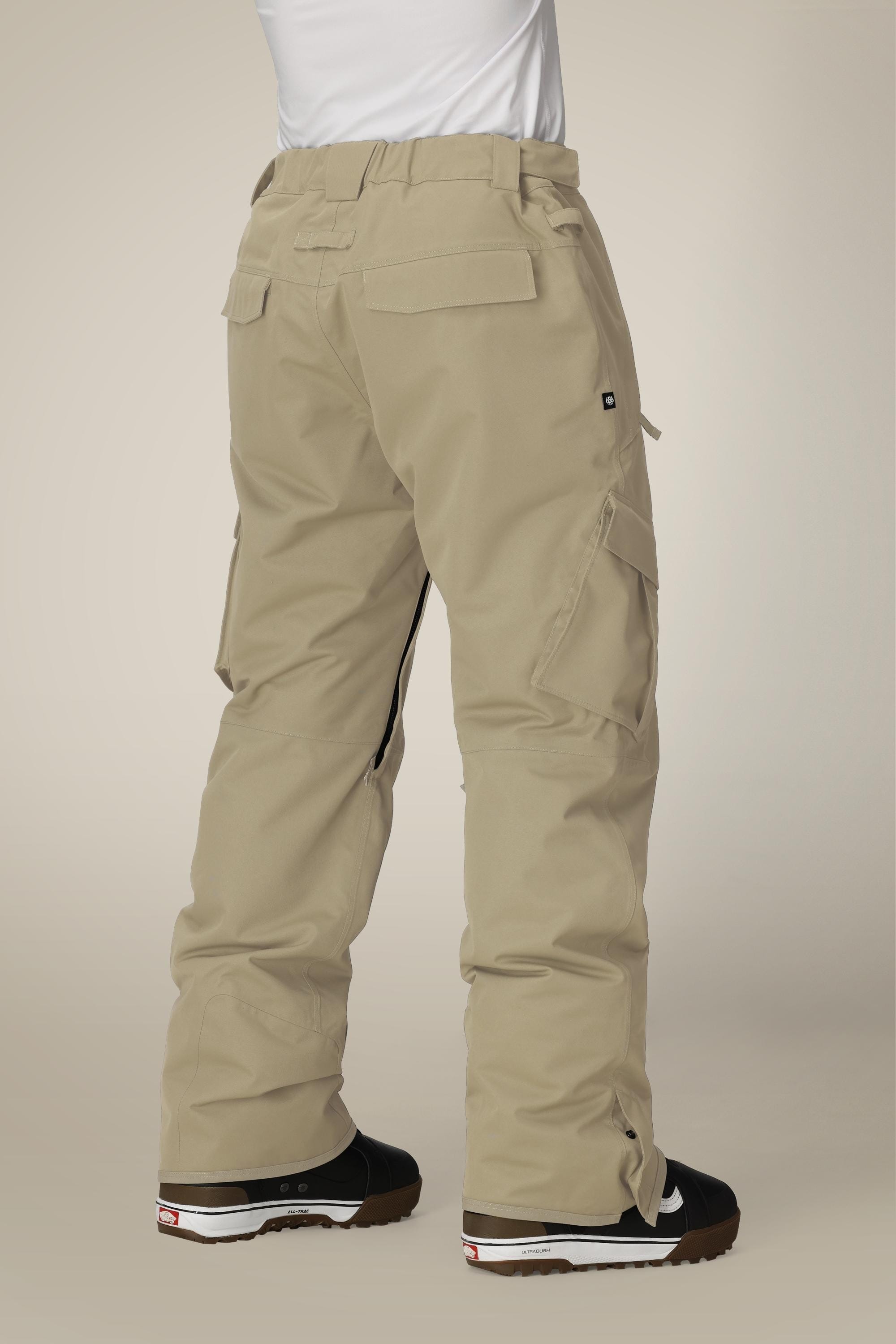 A person wearing 686 Mens Infinity Insulated Cargo Pant by 686 stands with their back to the camera. The beige, loose-fitting pants feature multiple pockets, gathered ankles, and are styled with black and white sneakers. Upper body is partly visible.