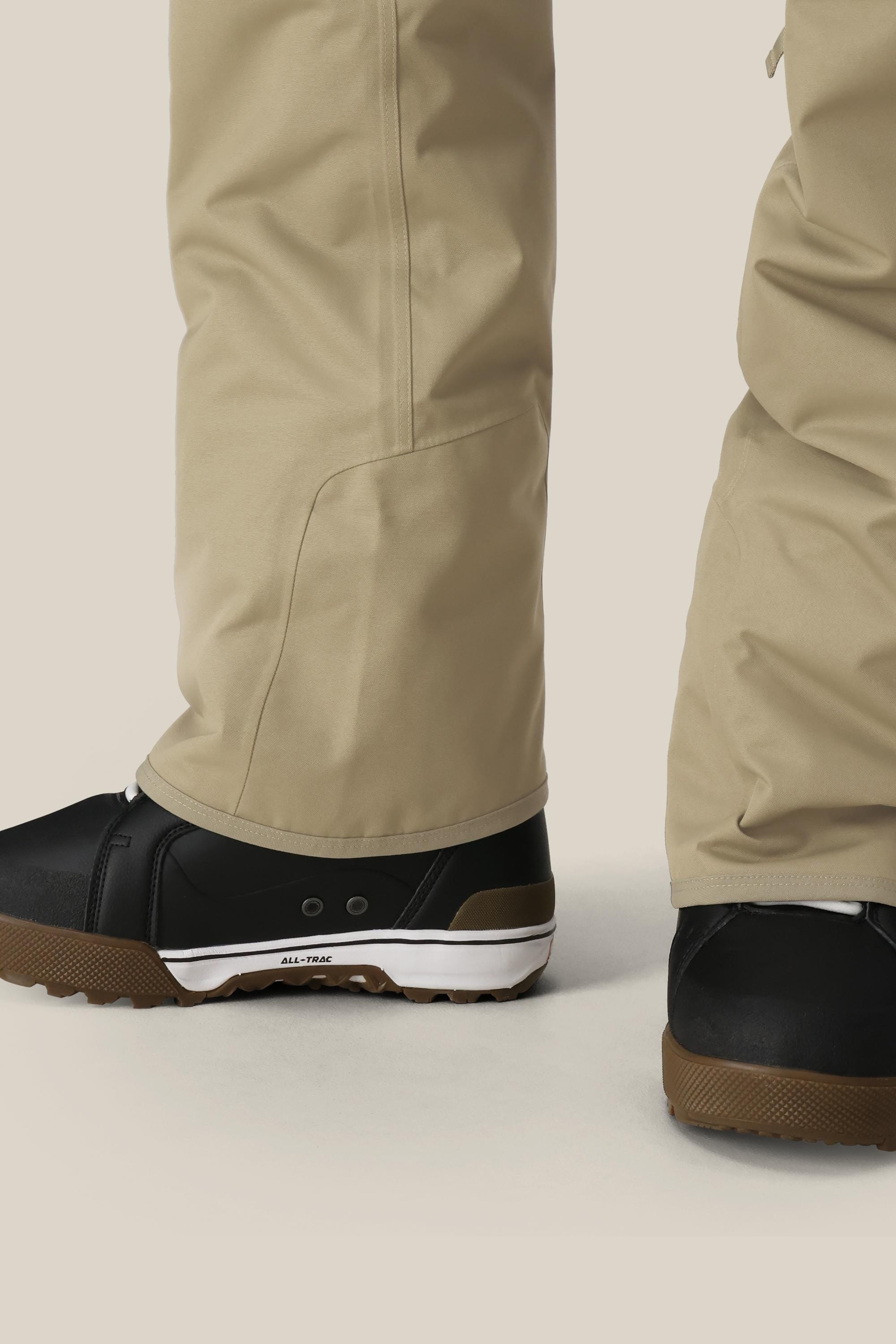 Close-up of a person wearing 686 Mens Infinity Insulated Cargo Pant by 686 in beige, paired with black shoes with brown and white soles. Only the lower legs and feet are visible on a light-colored surface.