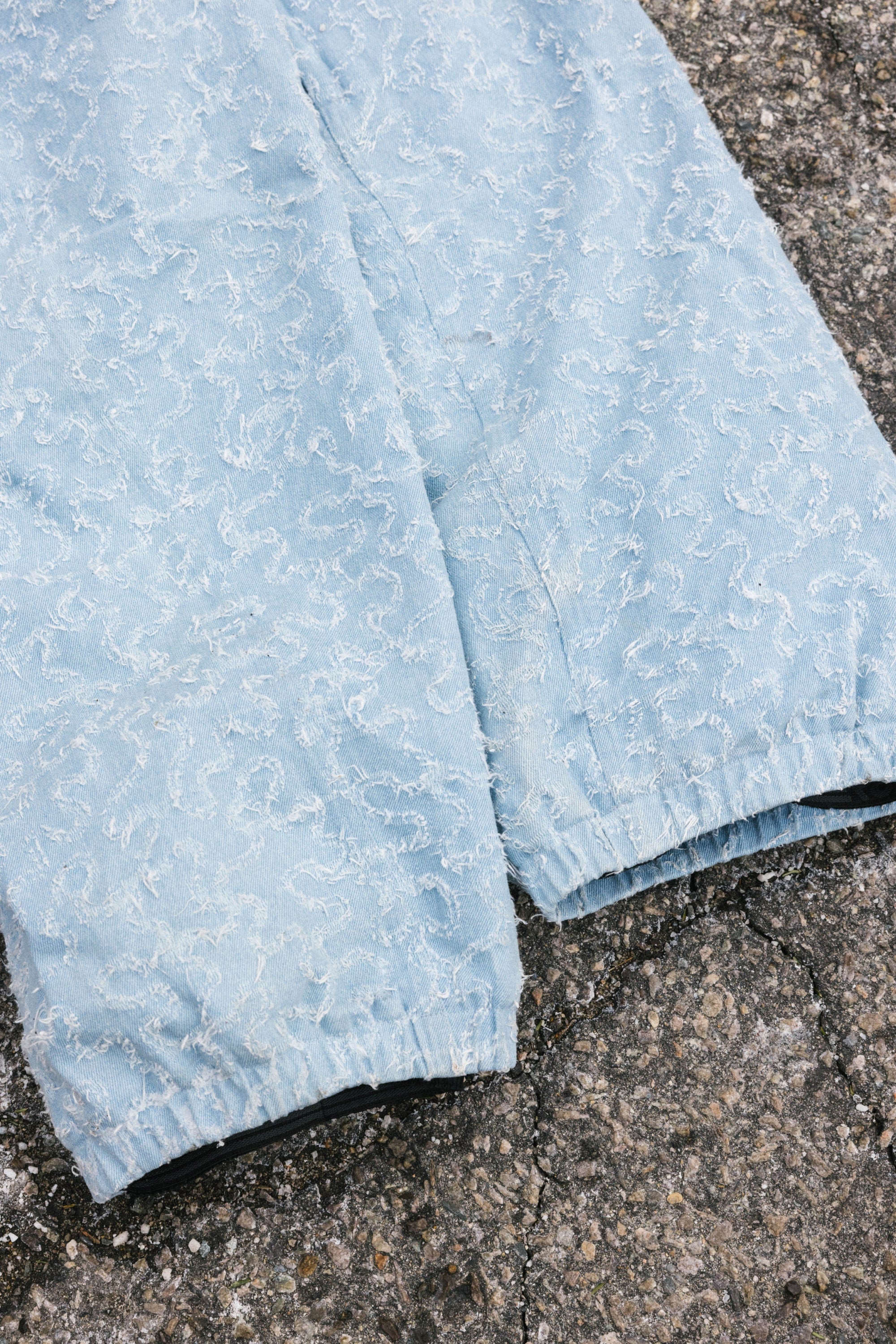 The 686 Dojo® Frayed Denim Pant by 686 features light blue men's woven denim with a subtle swirling texture and elastic cuffs, displayed on a rough, cracked concrete surface.