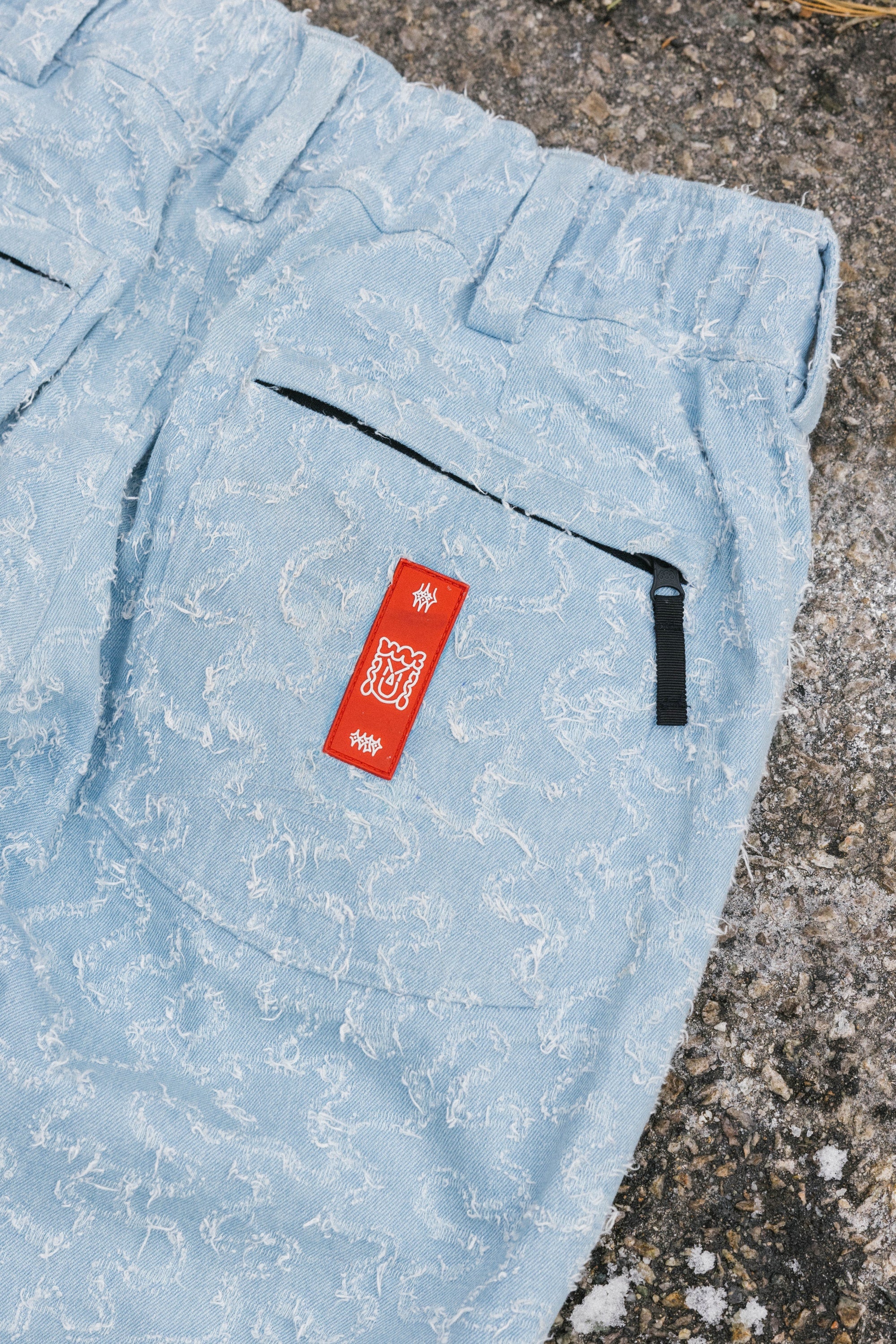 The 686 Dojo® Frayed Denim Pant by 686 is a light blue, textured men's woven pant with a subtle dragon pattern, black-zippered back pocket, and red tag with white symbols, displayed on rough gray concrete.