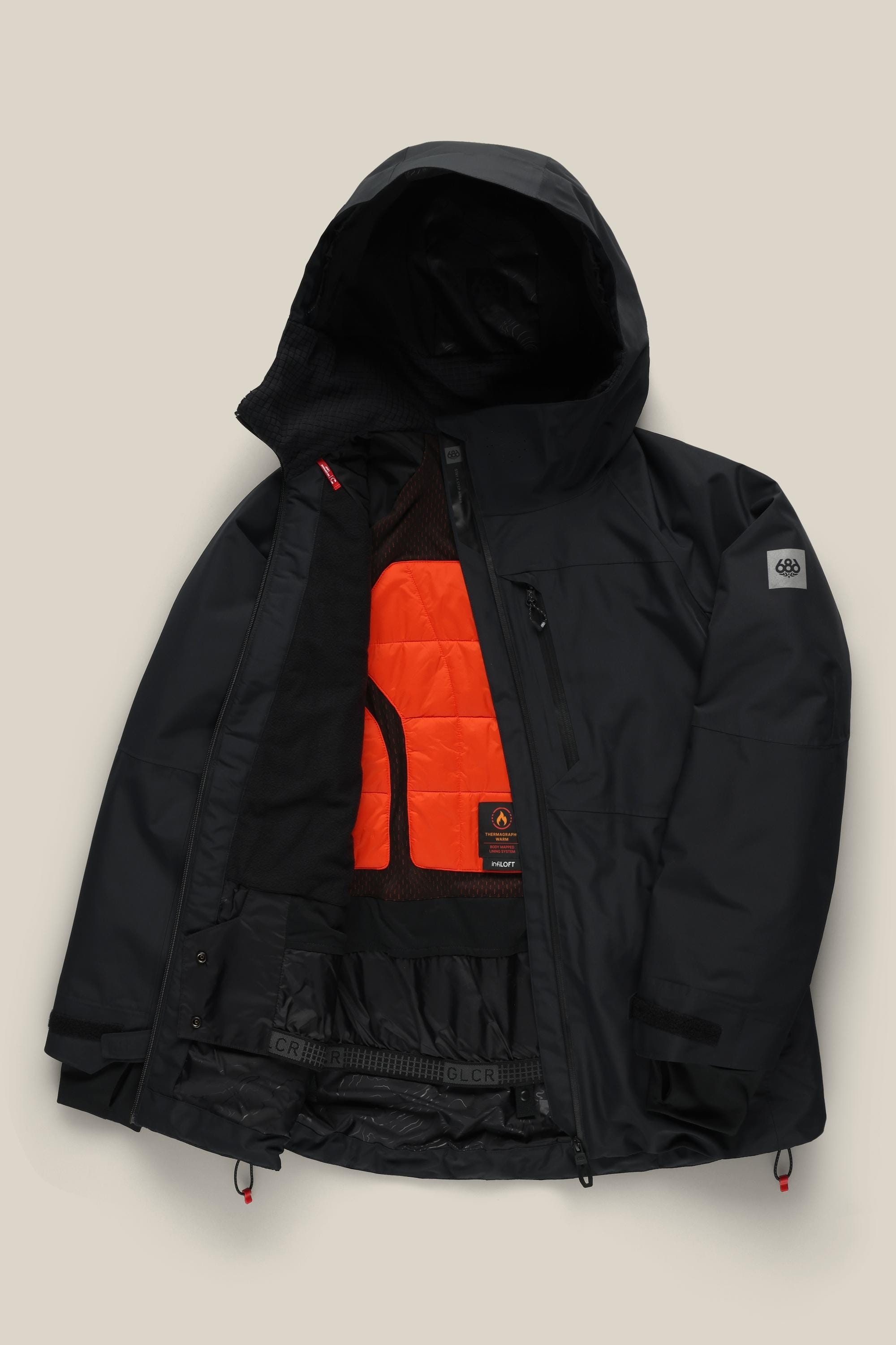The 686 Women’s Hydra™ Thermagraph® Jacket by 686 is a black hooded winter jacket with a partially open front that reveals an orange quilted inner lining and mesh pockets. It features zippered pockets and a logo patch on the sleeve.