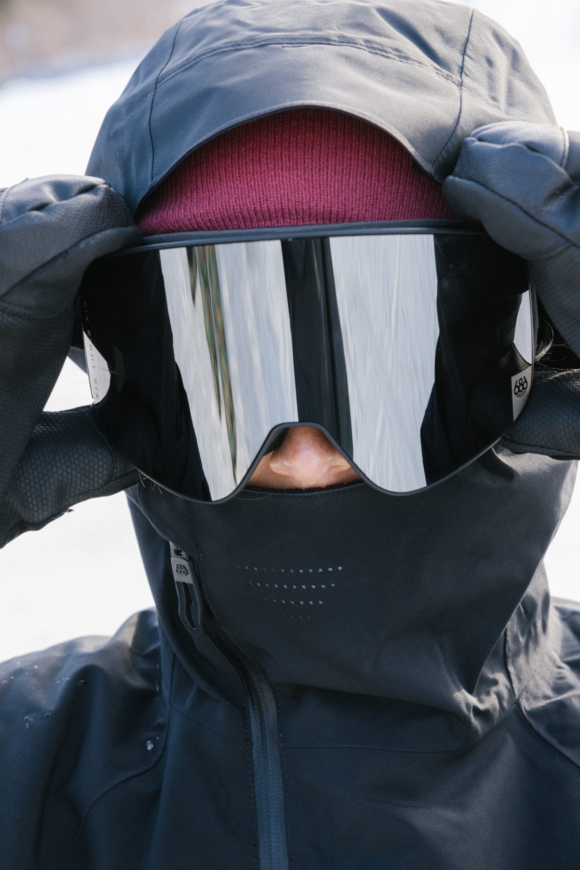 A person wearing a maroon beanie, reflective ski goggles, and the 686 Women’s Hydra™ Thermagraph® Jacket from 686 is adjusting their goggles outdoors in a snowy environment.