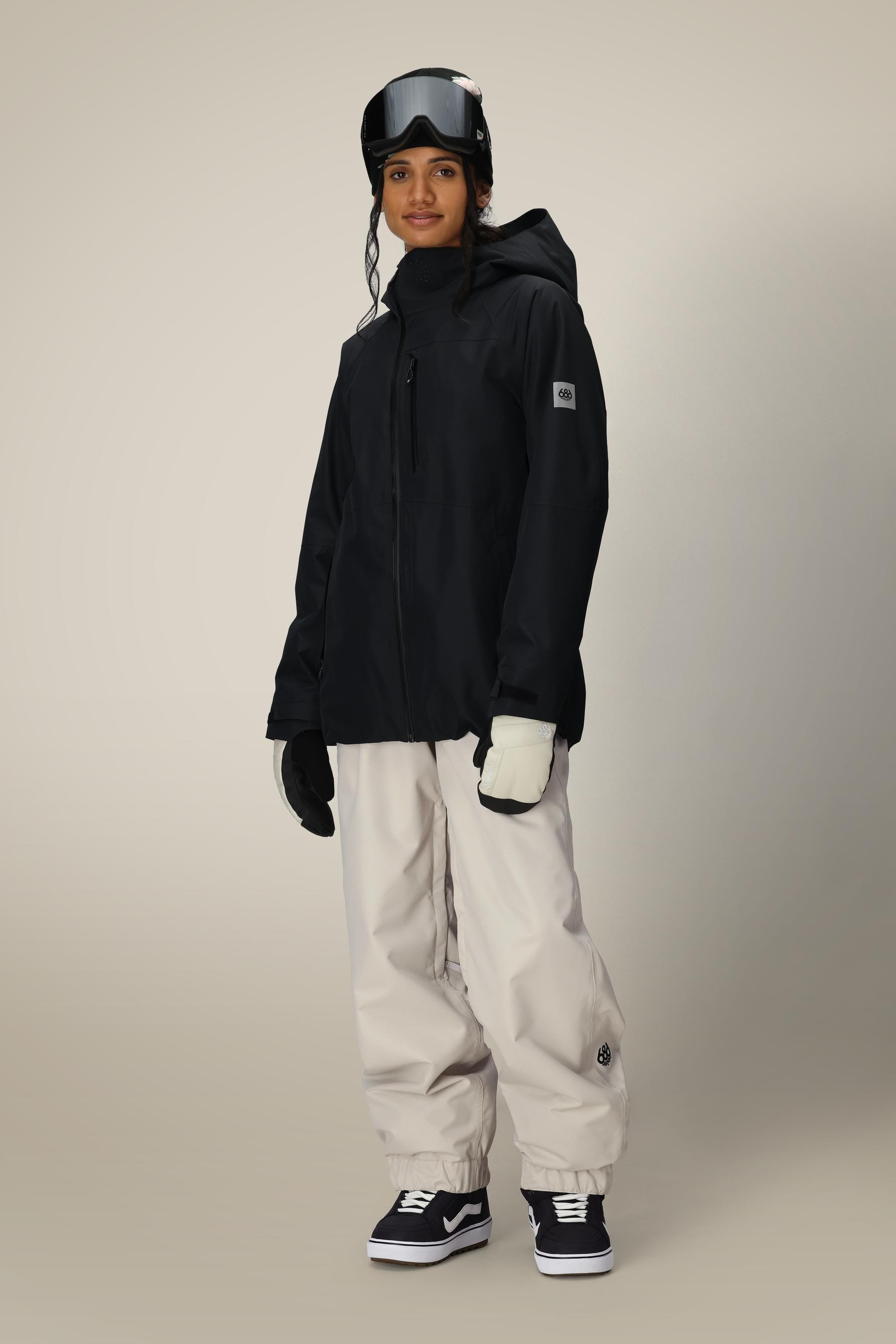 A person wearing the 686 Women’s Hydra™ Thermagraph® Jacket by 686, paired with white gloves, beige snow pants, black and white sneakers, and ski goggles, stands against a plain background looking ready for winter sports.