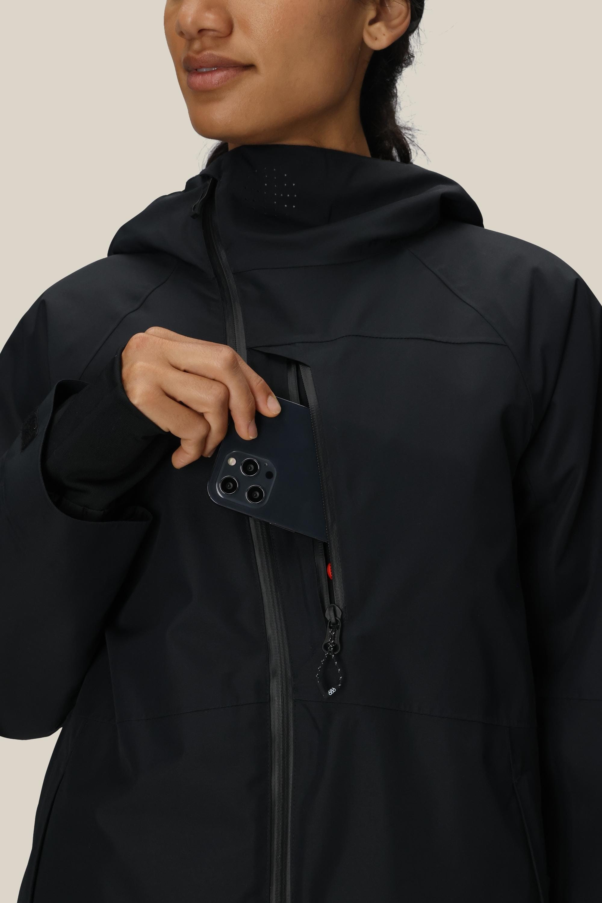 A woman wearing the 686 Women’s Hydra™ Thermagraph® Jacket is placing a black smartphone into the jacket’s zippered chest pocket. She is visible from the shoulders up and is gazing off to the side with a slight smile.
