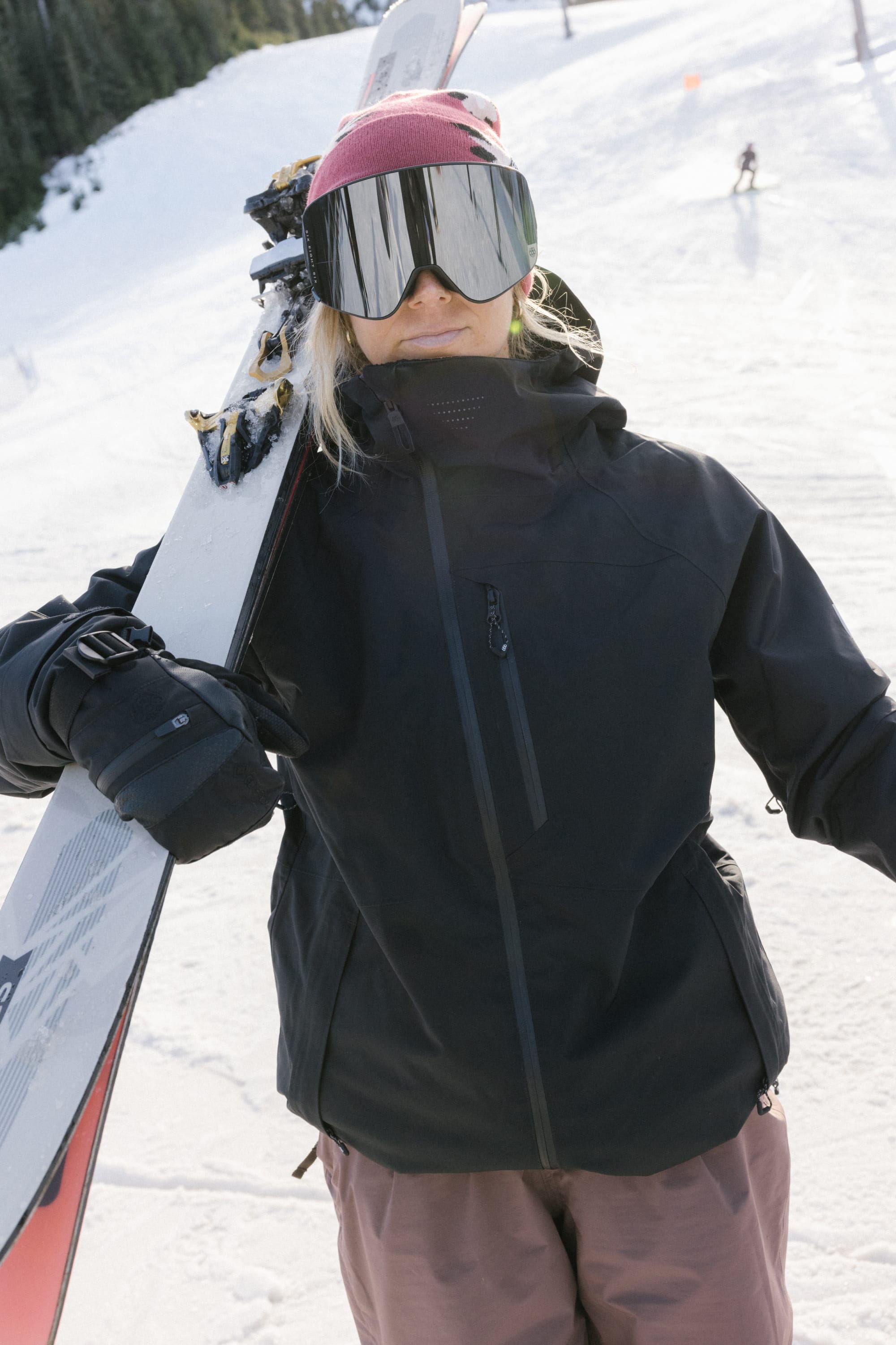 Wearing the 686 Women’s Hydra™ Thermagraph® Jacket, a person with a pink beanie and large reflective ski goggles holds skis on their shoulder while standing on a snowy slope with trees and other skiers in the background.