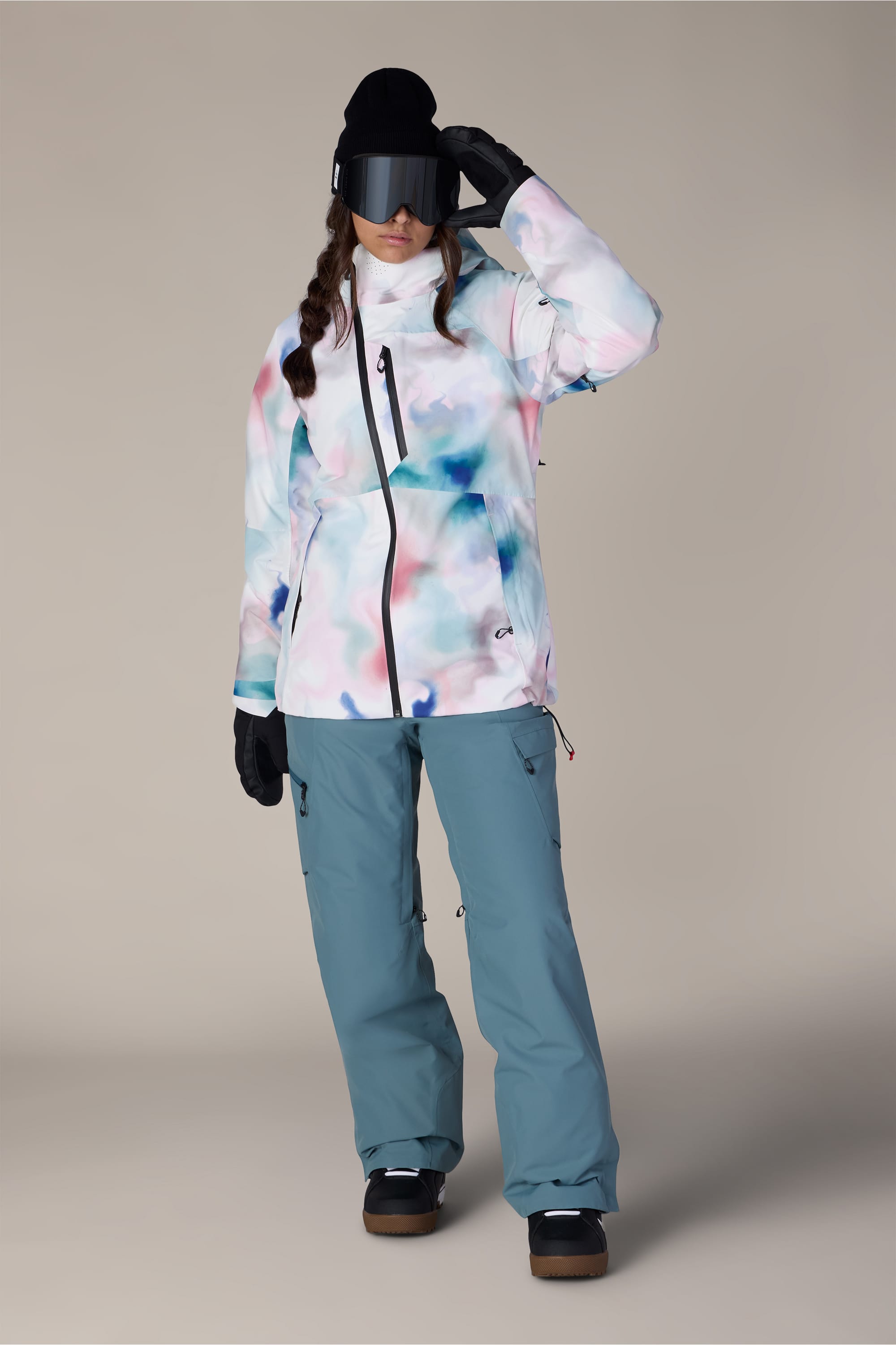 A person wearing the 686 Women’s Hydra™ Thermagraph® Jacket by 686 stands against a plain background, paired with blue snow pants, black gloves, black boots, a black beanie, and large black ski goggles.