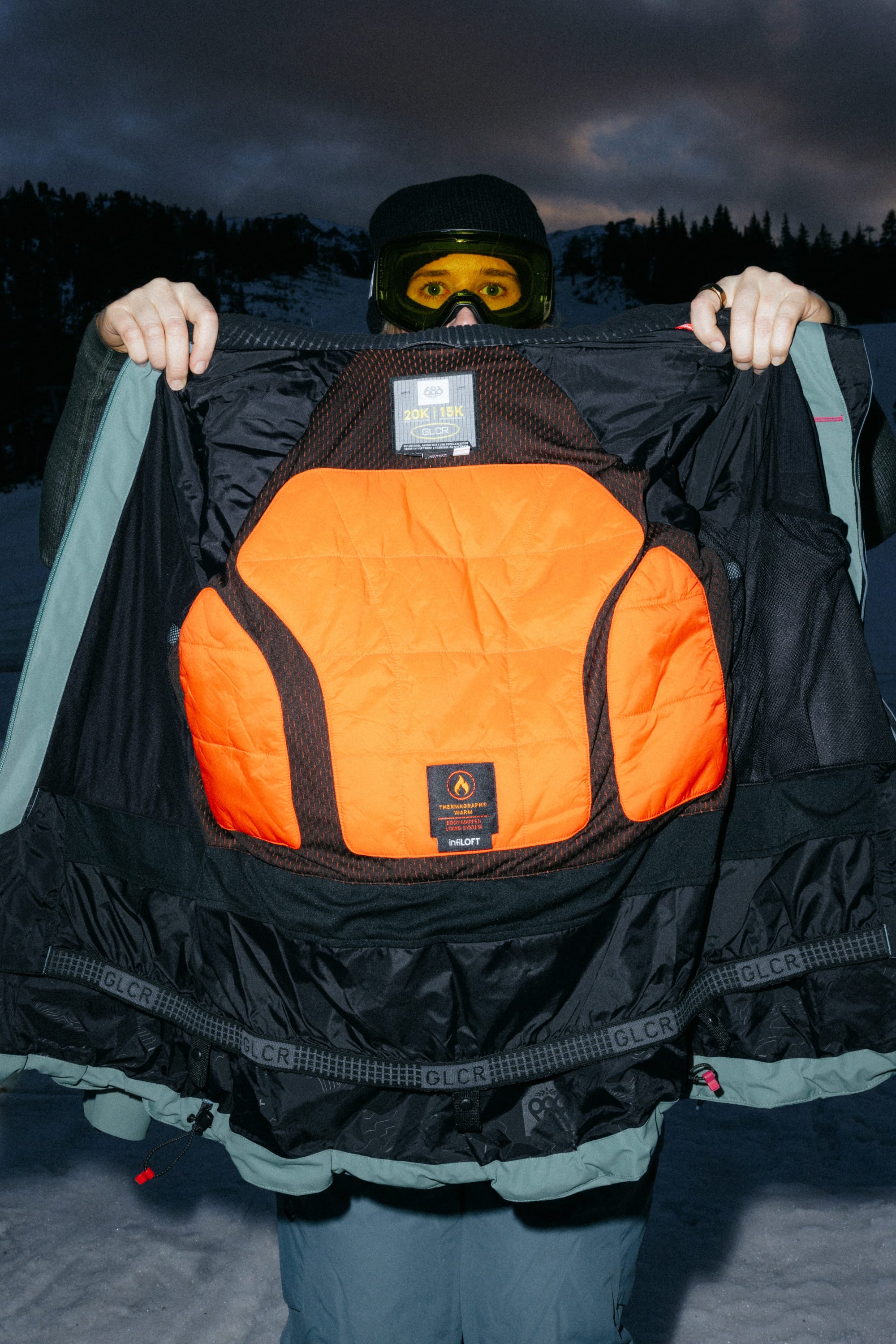 Wearing the 686 Women’s Hydra™ Thermagraph® Jacket, a person in snow gear opens their coat to reveal its vibrant orange padded interior, with snow and trees in the background beneath a moody, cloudy sky.