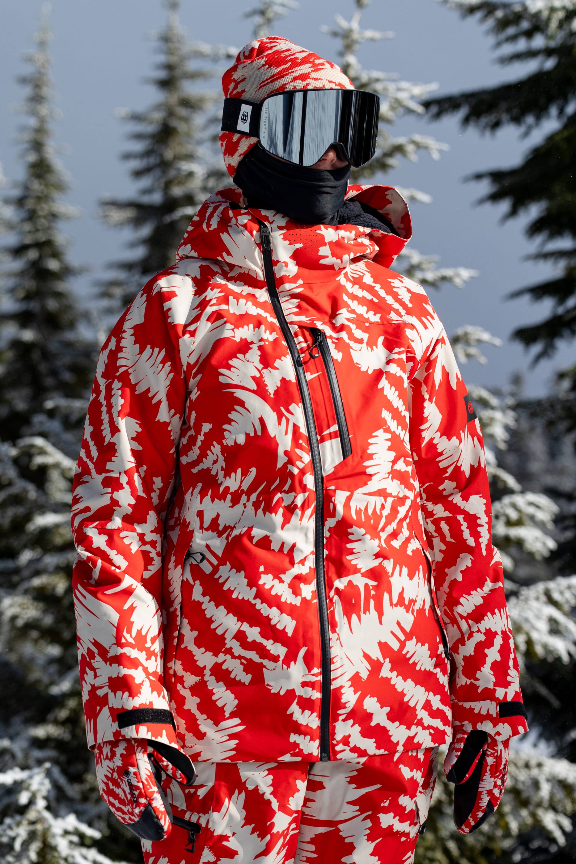 Wearing the 686 Women’s Hydra™ Thermagraph® Jacket by 686, a person with mirrored ski goggles and a hood stands outdoors in a snowy forest with evergreen trees in the background.