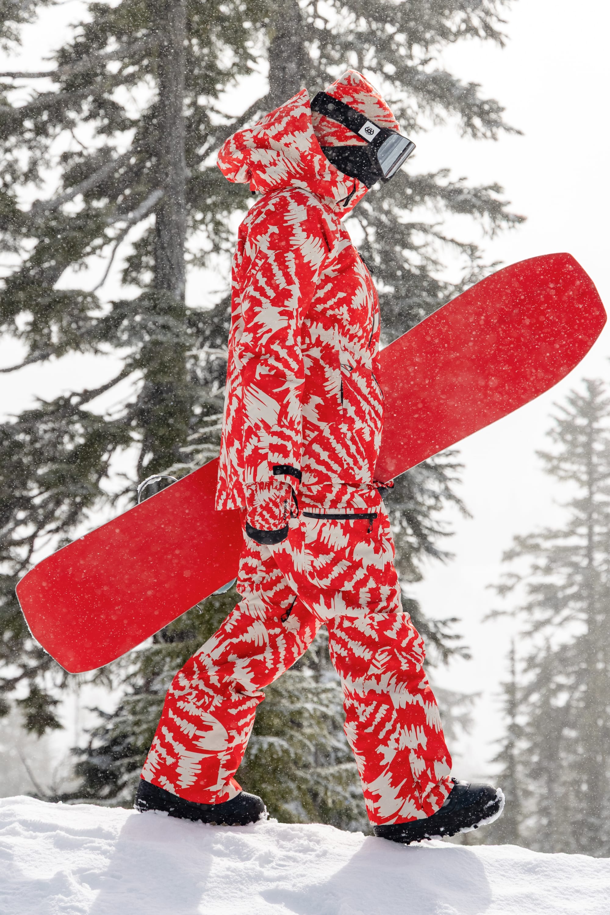 Wearing the 686 Women’s Hydra™ Thermagraph® Jacket, a person walks through the snow holding a red snowboard, with snow-covered trees and falling snow in the background.
