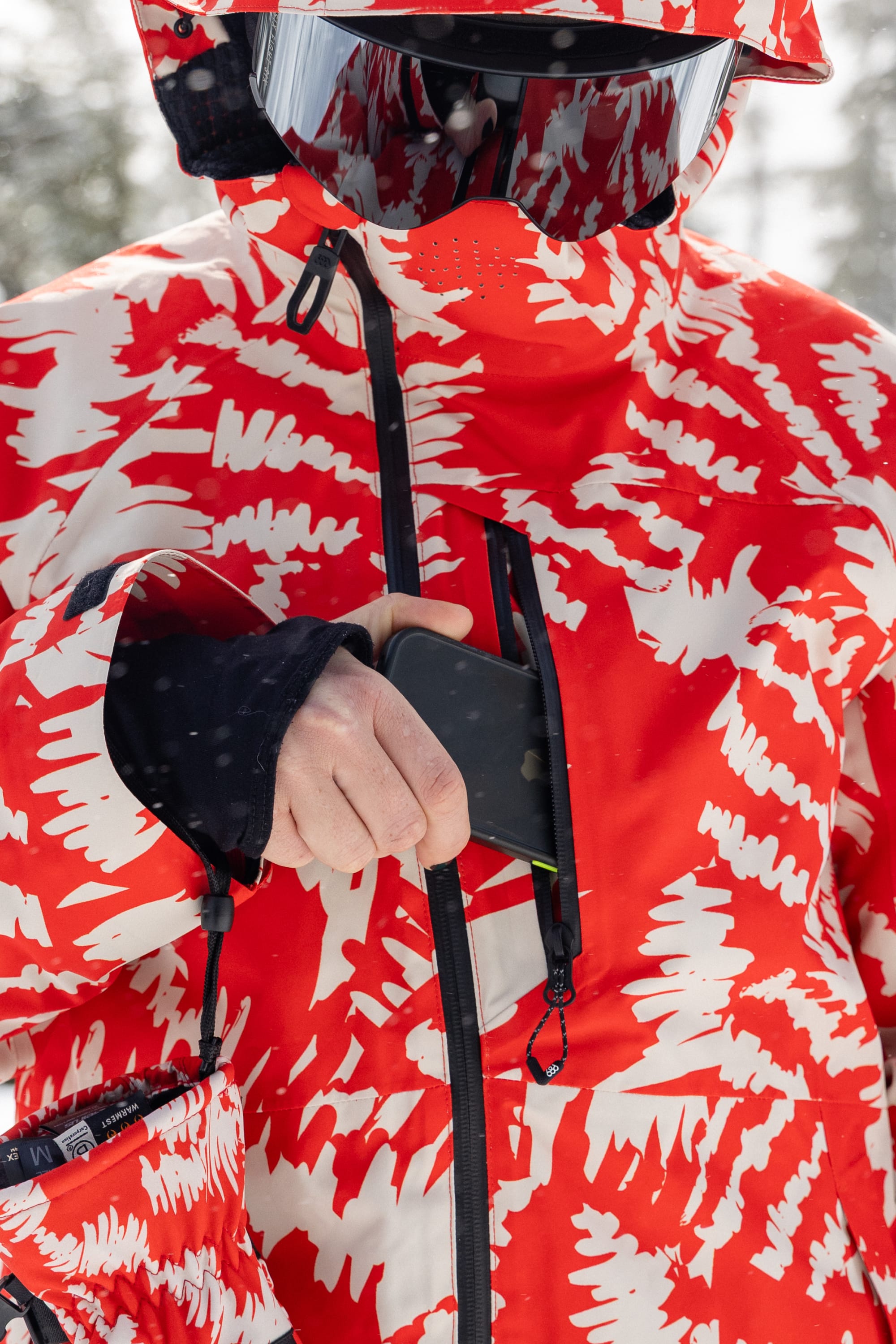 A person wearing the 686 Women’s Hydra™ Thermagraph® Jacket with a red and white pattern and goggles unzips a chest pocket to retrieve or store a smartphone, while snowflakes are visible on the jacket.