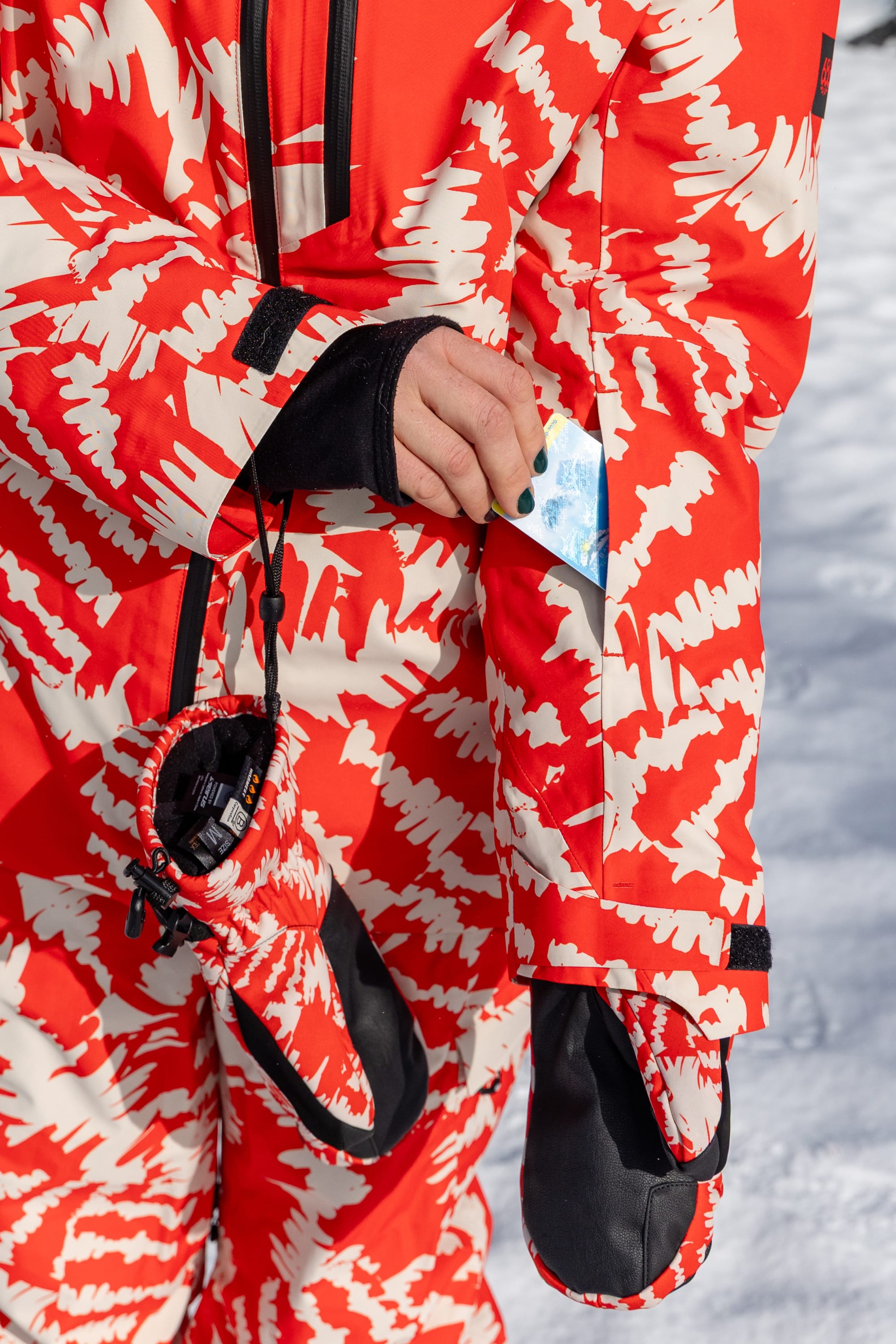 Wearing the 686 Women’s Hydra™ Thermagraph® Jacket, a person stands on snow, placing an item into a zippered sleeve pocket as a matching glove from 686 dangles from their wrist.