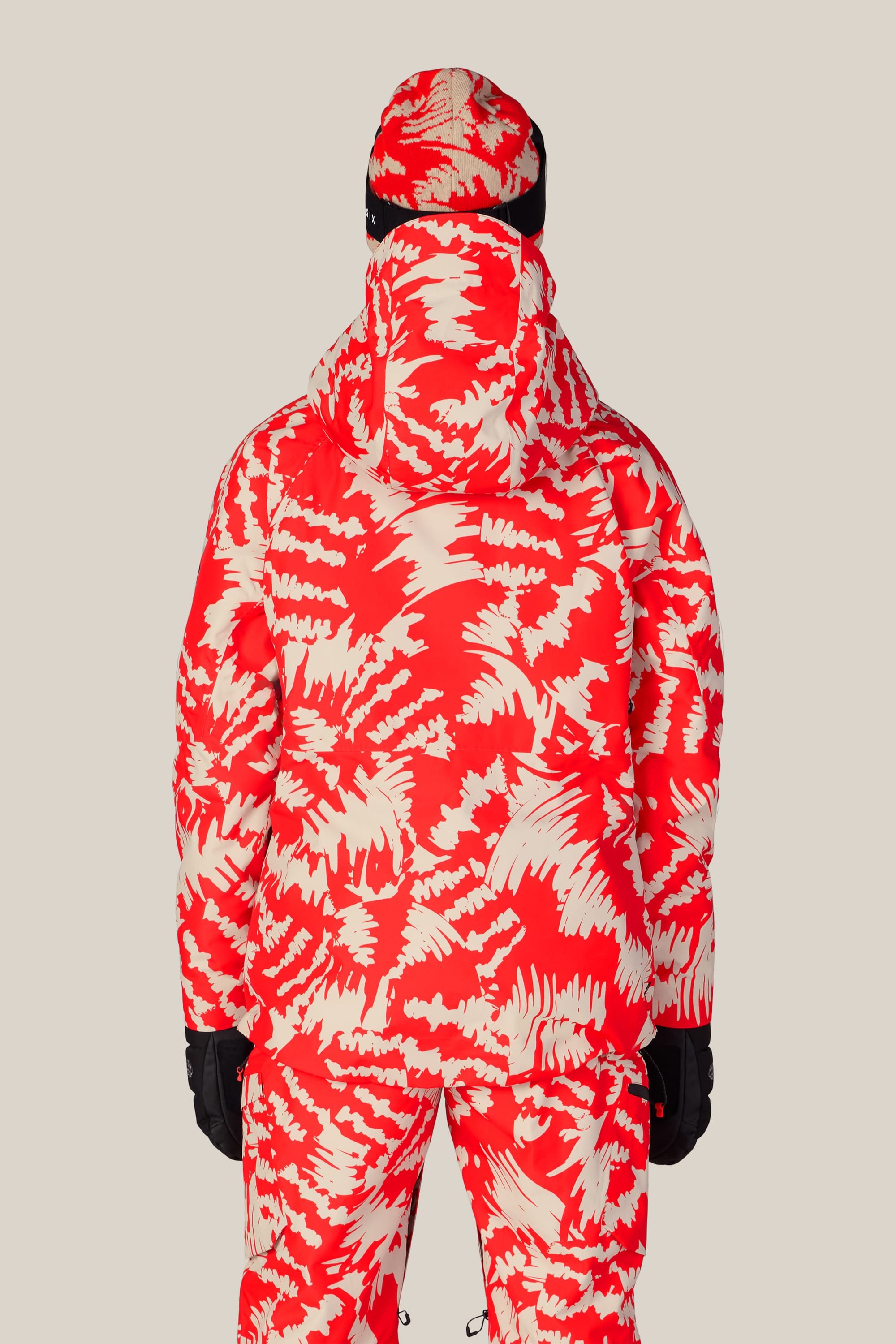 A person stands facing away, wearing the 686 Women’s Hydra™ Thermagraph® Jacket in a red and beige abstract pattern, along with matching winter pants, gloves, and hat by 686, set against a plain light background.