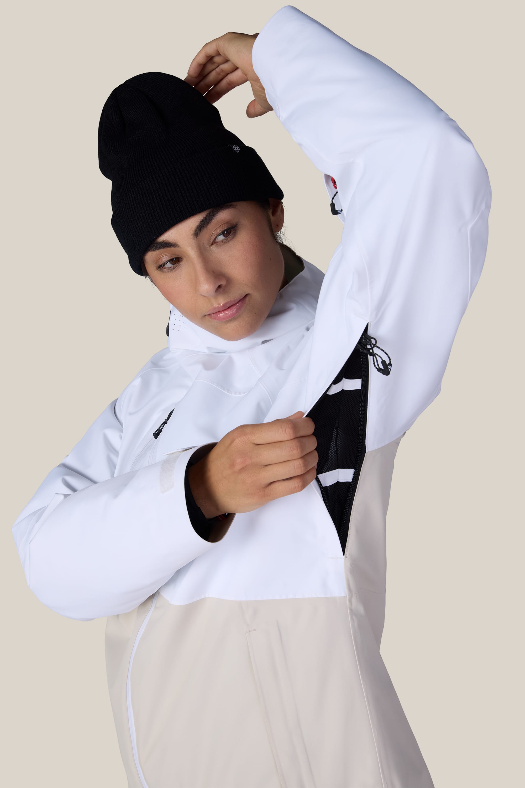 A woman wearing the 686 Women’s Hydra™ Thermagraph® Jacket in white and a black beanie demonstrates the underarm ventilation zipper, standing against a plain light background.