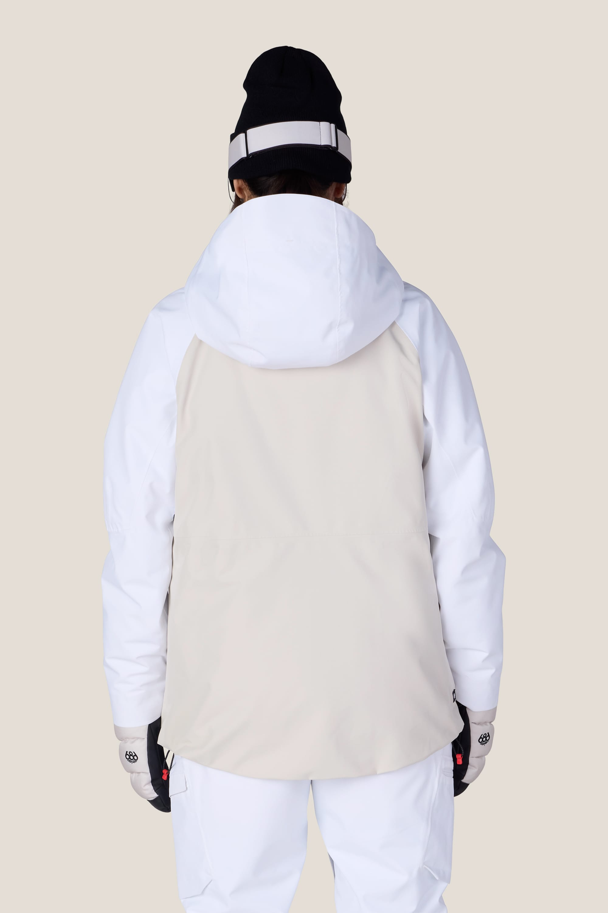 A person faces away, wearing the 686 Women’s Hydra™ Thermagraph® Jacket in white and beige, paired with white pants, a black beanie, and gloves against a plain light-colored background.