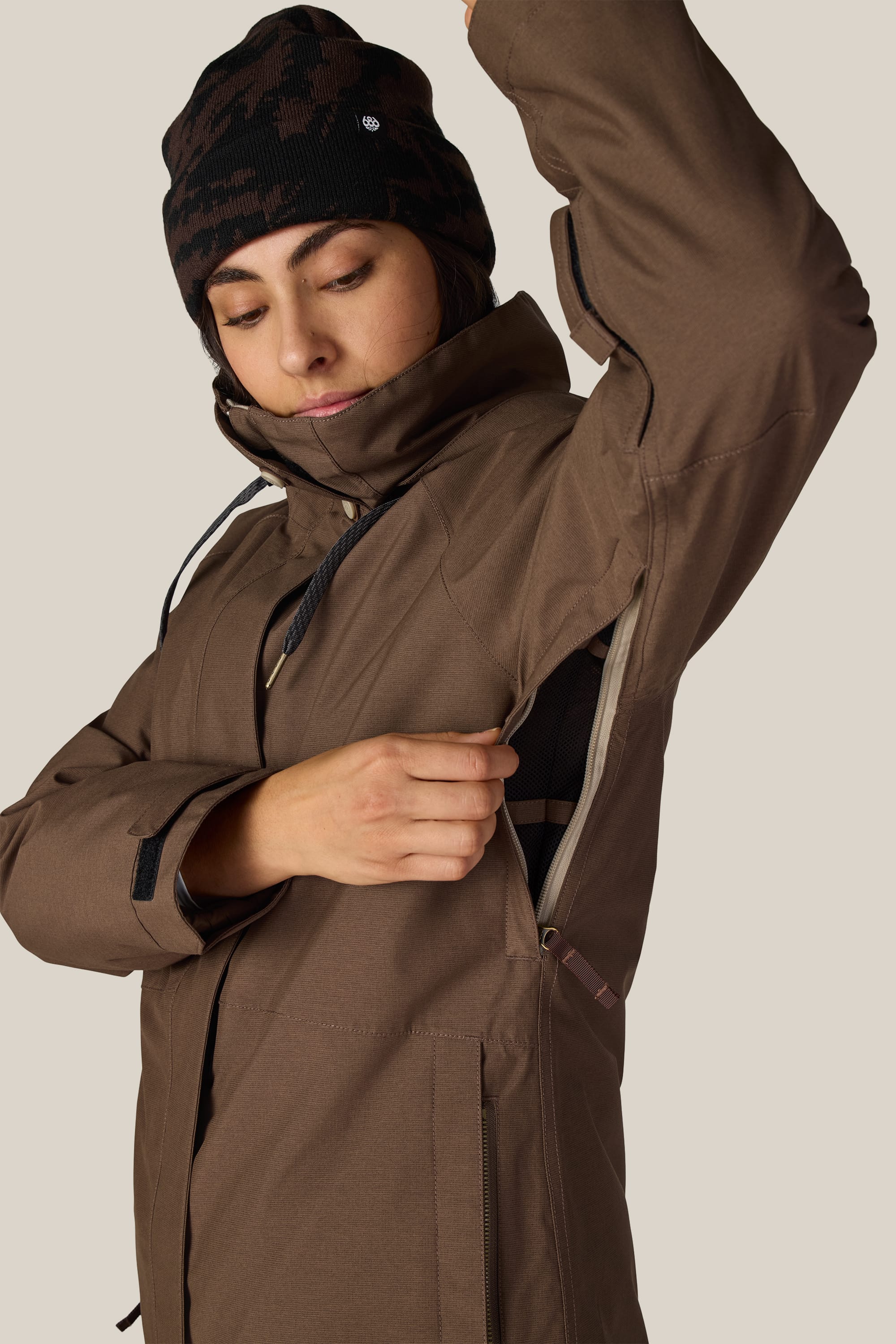 A person wearing the 686 Women’s SMARTY® 3-In-1 Spellbound Jacket in brown and a black beanie unzips the jacket’s underarm ventilation. The background is plain and light-colored.