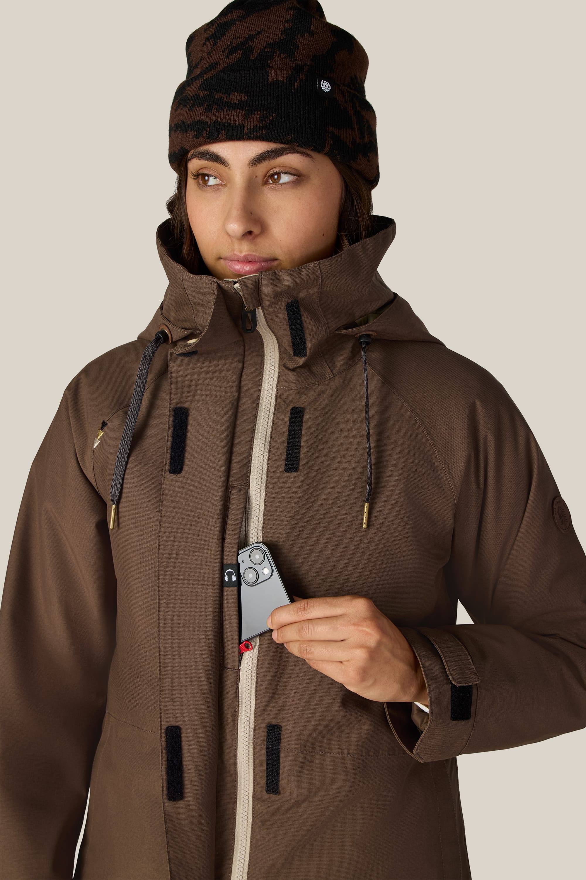 Wearing the 686 Women’s SMARTY® 3-In-1 Spellbound Jacket and a patterned beanie, a person places a phone into the jacket’s chest pocket, glancing slightly to the side against a plain background.