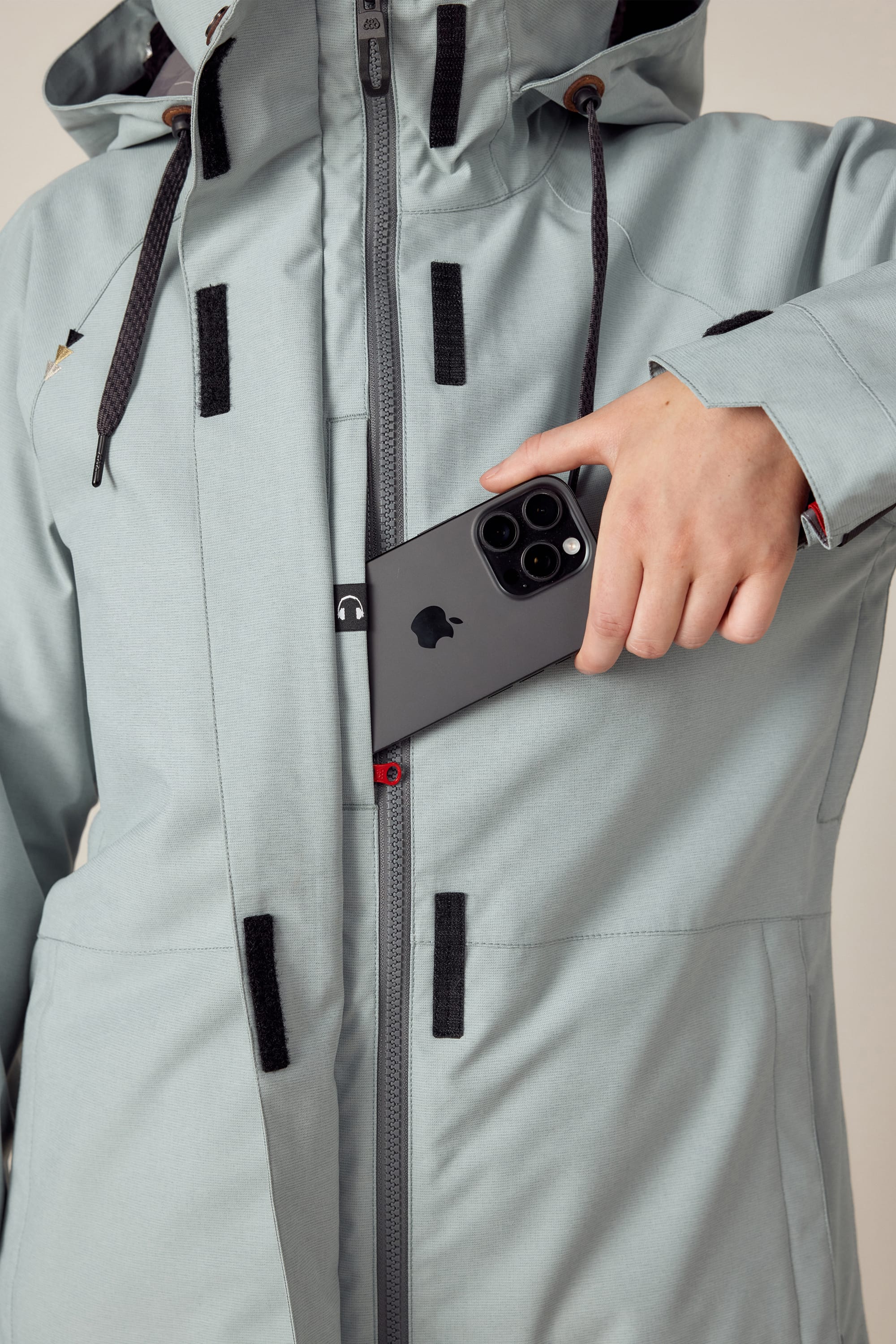 A woman wearing the 686 Womens Smarty 3-In-1 Spellbound Jacket in light gray slips a black smartphone with three cameras into the chest pocket, which is partially unzipped to reveal the phone. The image is cropped to show only her torso and hand.