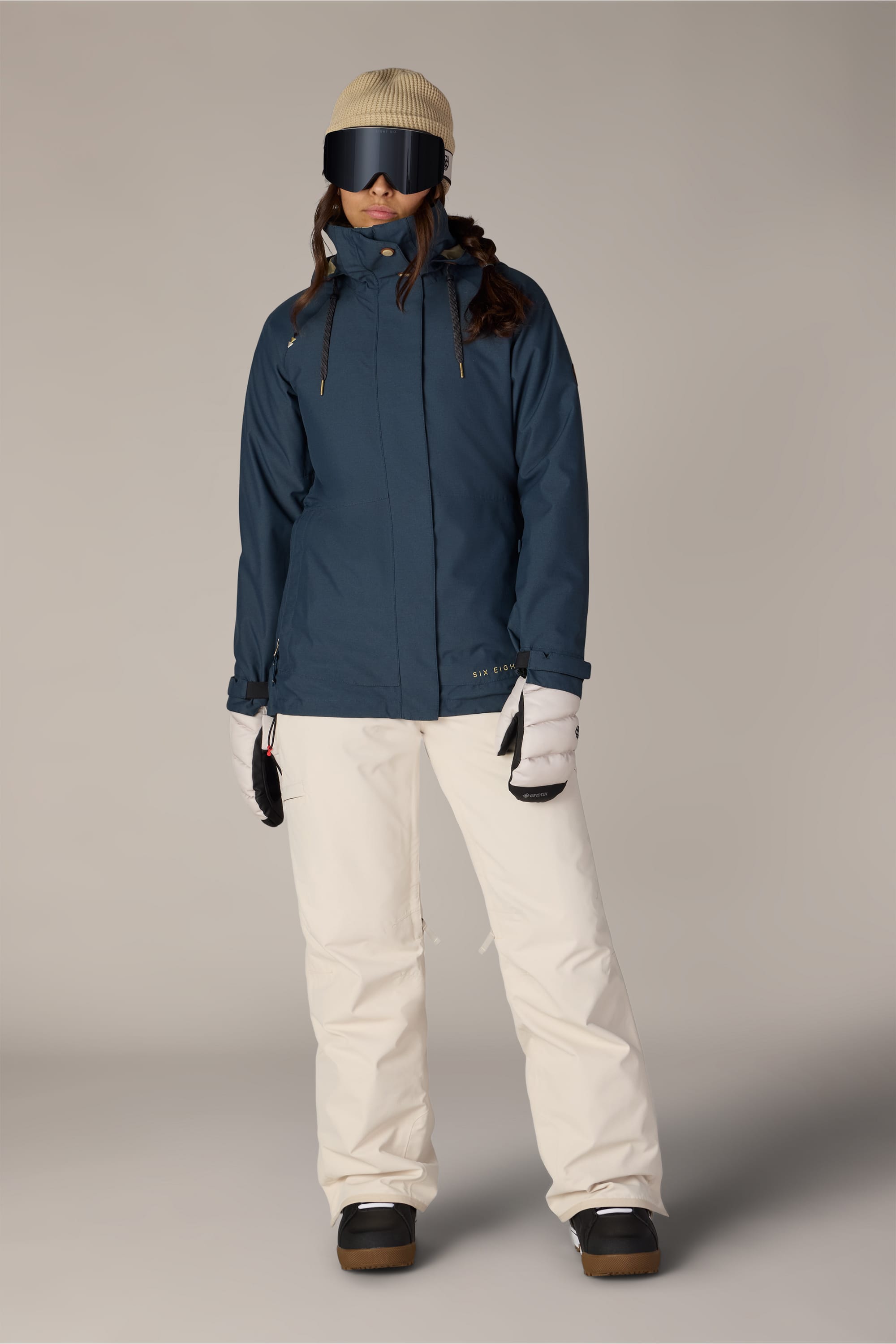 A person wearing the 686 Women’s SMARTY® 3-In-1 Spellbound Jacket stands facing forward against a plain background, styled with white snow pants, white gloves, black goggles, a beige beanie, and black boots.