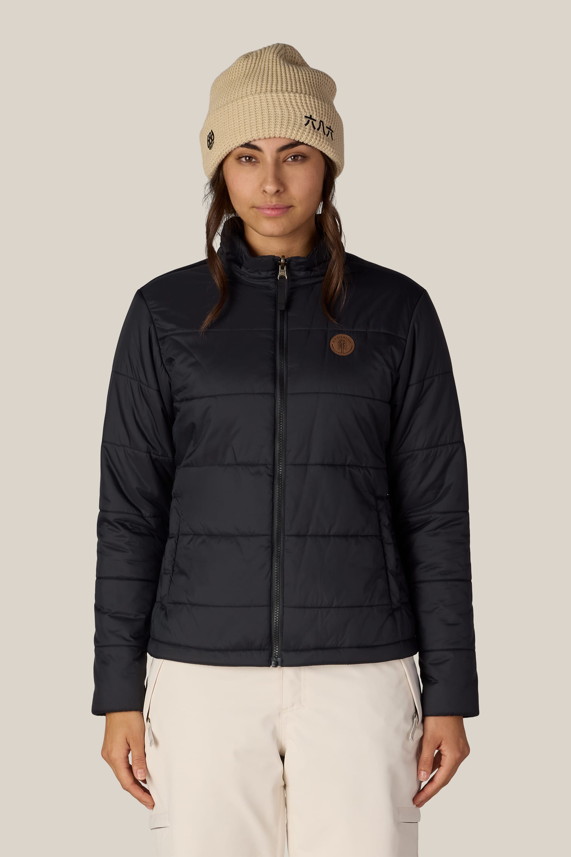 A person wearing the 686 Women’s SMARTY® 3-In-1 Spellbound Jacket in black, along with a beige knit beanie and light-colored pants, stands facing forward against a plain beige background.