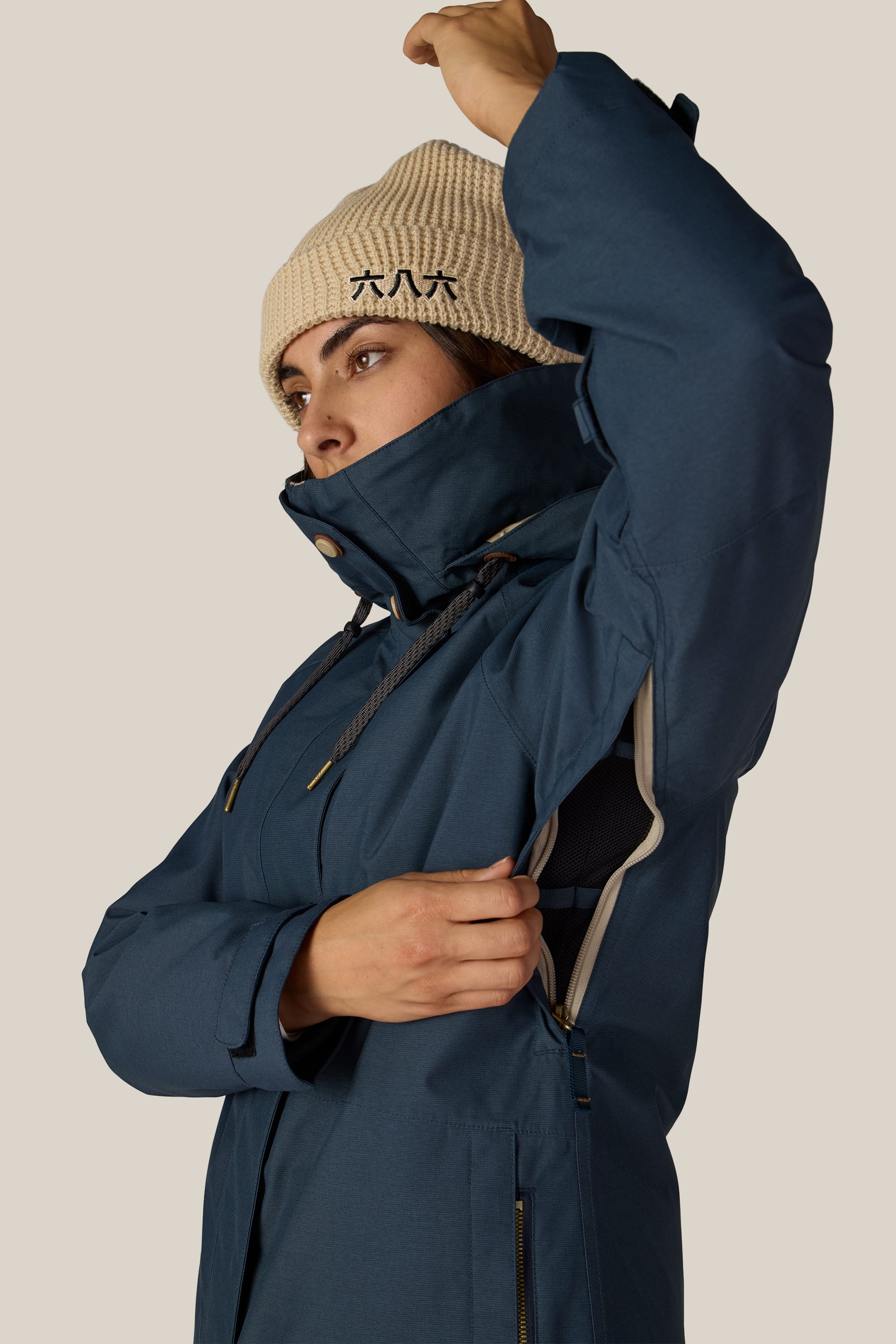 Wearing the 686 Women’s SMARTY® 3-In-1 Spellbound Jacket and a beige beanie, a person opens the underarm zipper of the jacket to reveal its mesh lining, posed against a plain background.