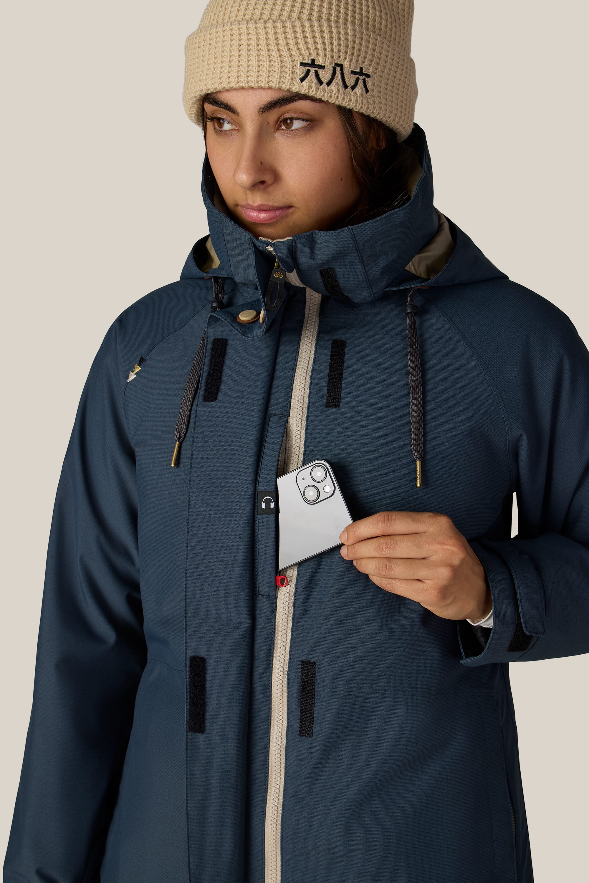 A woman wearing the 686 Women’s SMARTY® 3-In-1 Spellbound Jacket in blue and a beige knit hat puts a silver smartphone into the chest pocket, glancing to the side.