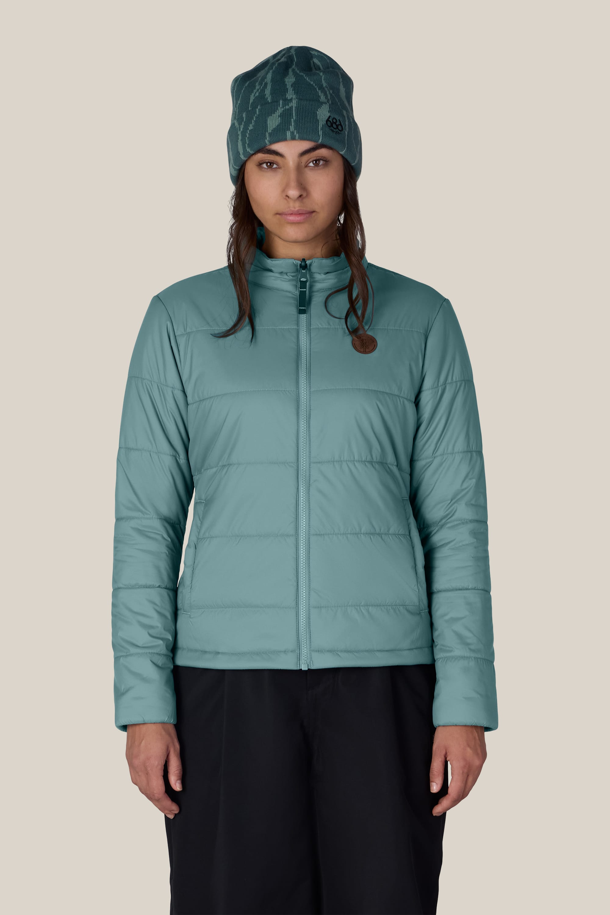 A woman wearing the 686 Women’s SMARTY® 3-In-1 Spellbound Jacket by 686, paired with black pants and a teal beanie, stands facing forward against a plain beige background.