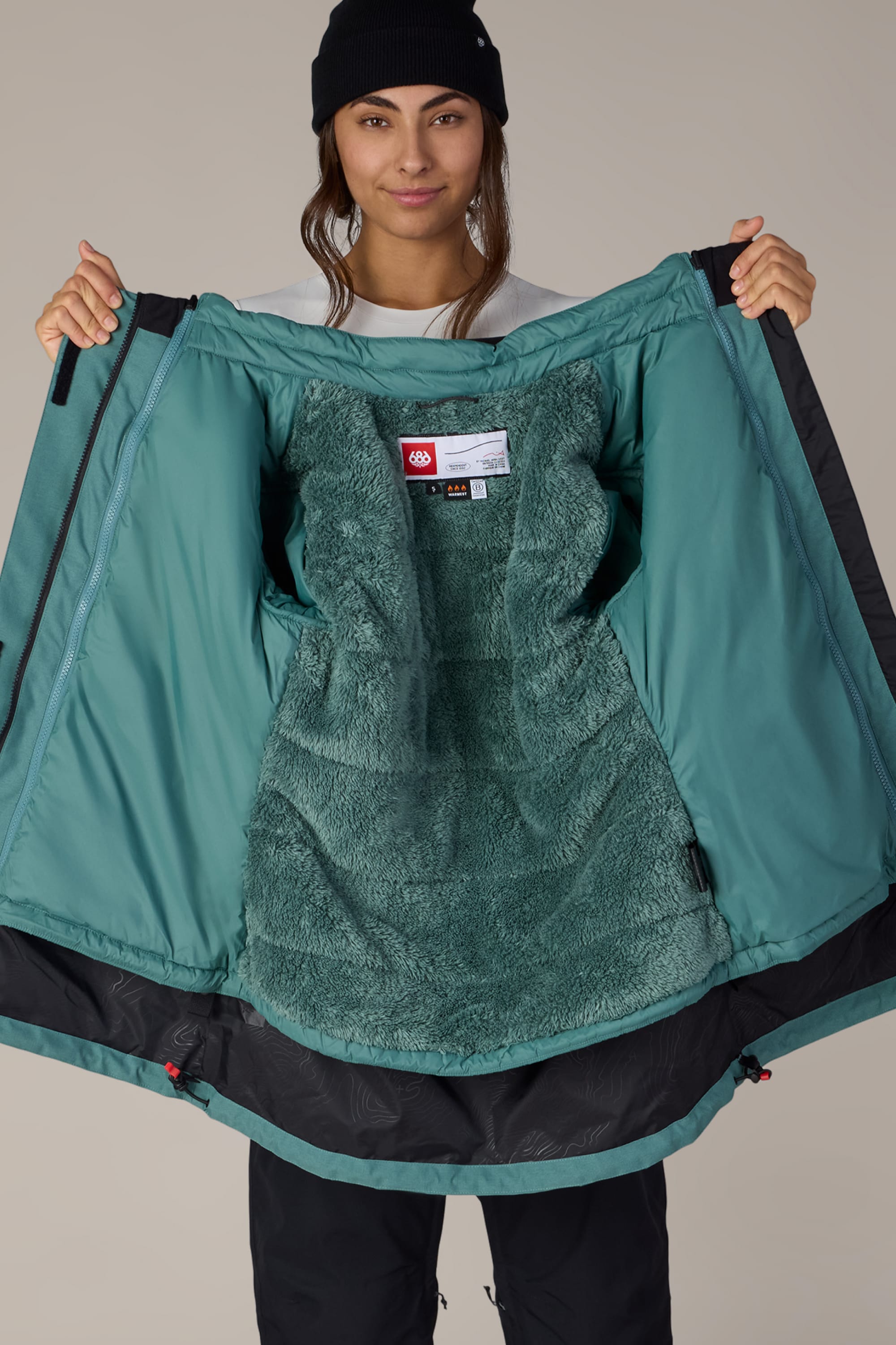 A person in a black beanie holds open the 686 Women’s SMARTY® 3-In-1 Spellbound Jacket, revealing its soft, fuzzy lining. The inner back of the green winter jacket features a 686 label, and the person smiles slightly at the camera.