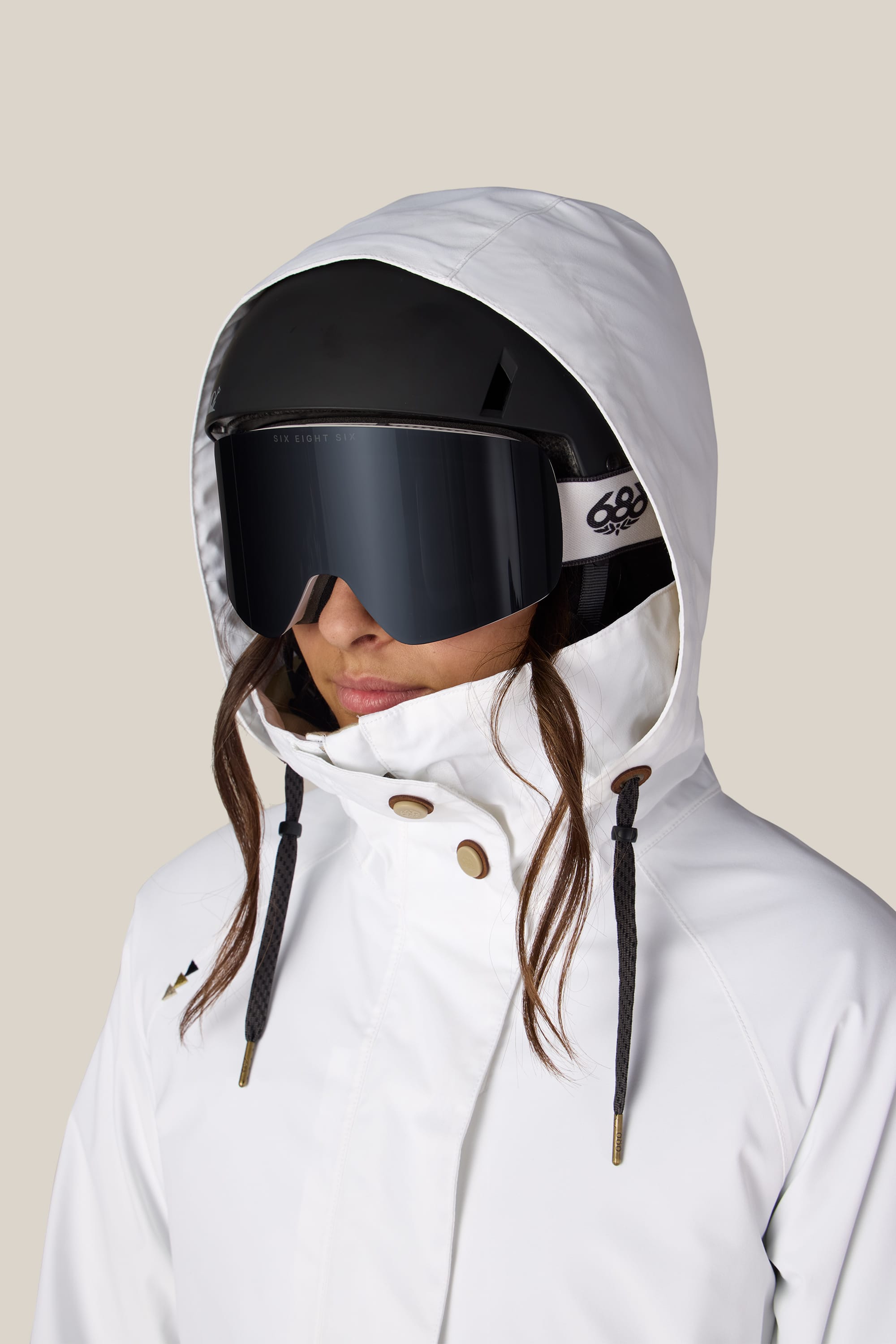 A person is wearing the 686 Women’s SMARTY® 3-In-1 Spellbound Jacket in white, paired with large black ski goggles and a black helmet, facing slightly to the side against a plain background.
