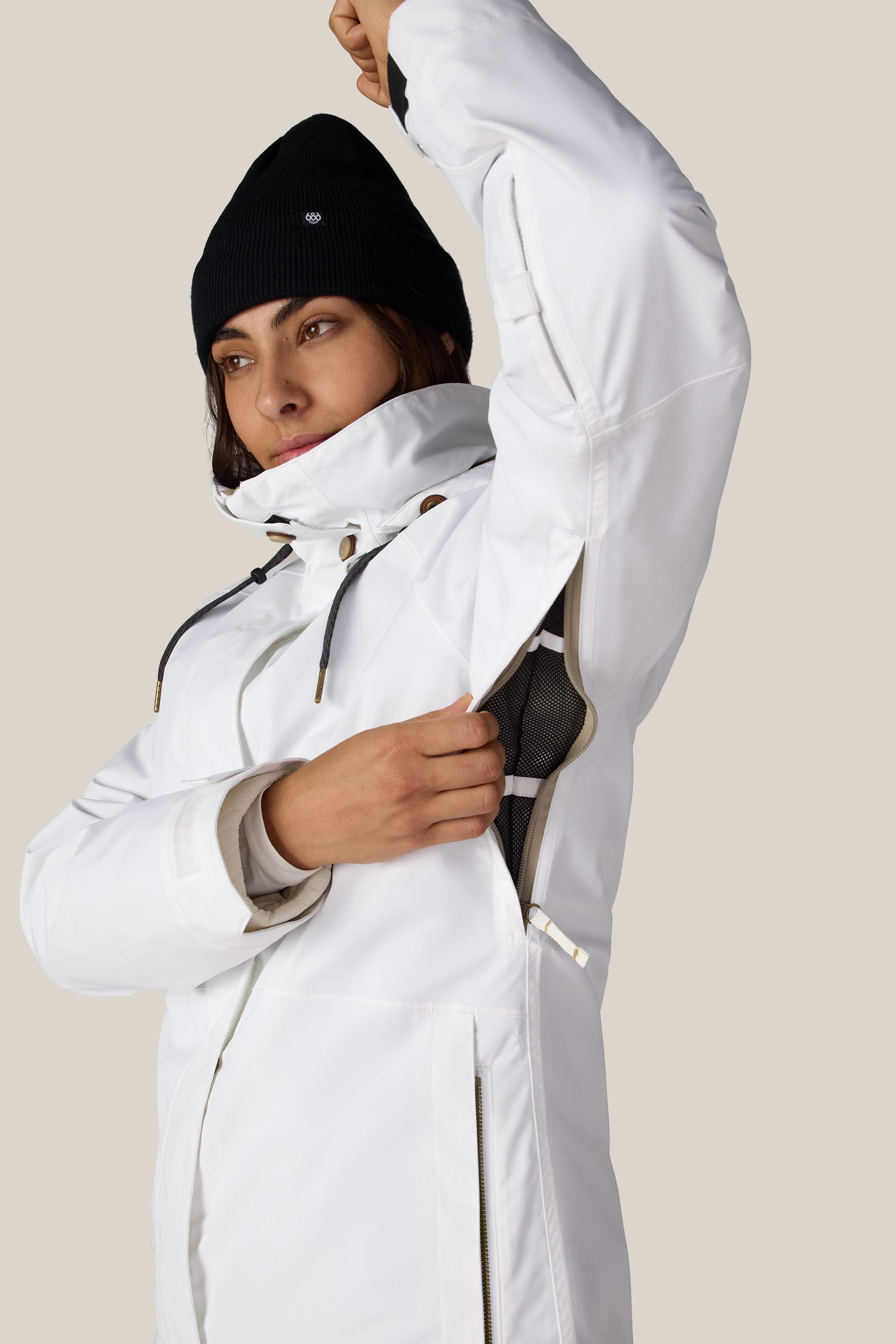 A woman wearing the 686 Women’s SMARTY® 3-In-1 Spellbound Jacket in white and a black beanie unzips an underarm vent against a plain, light-colored background.
