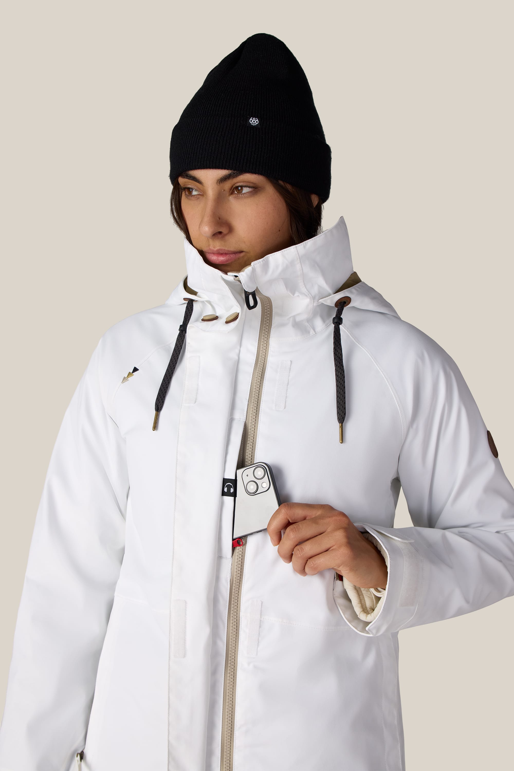A person wearing the 686 Women’s SMARTY® 3-In-1 Spellbound Jacket in white and a black beanie holds a phone with a triple camera partially inside their jacket.