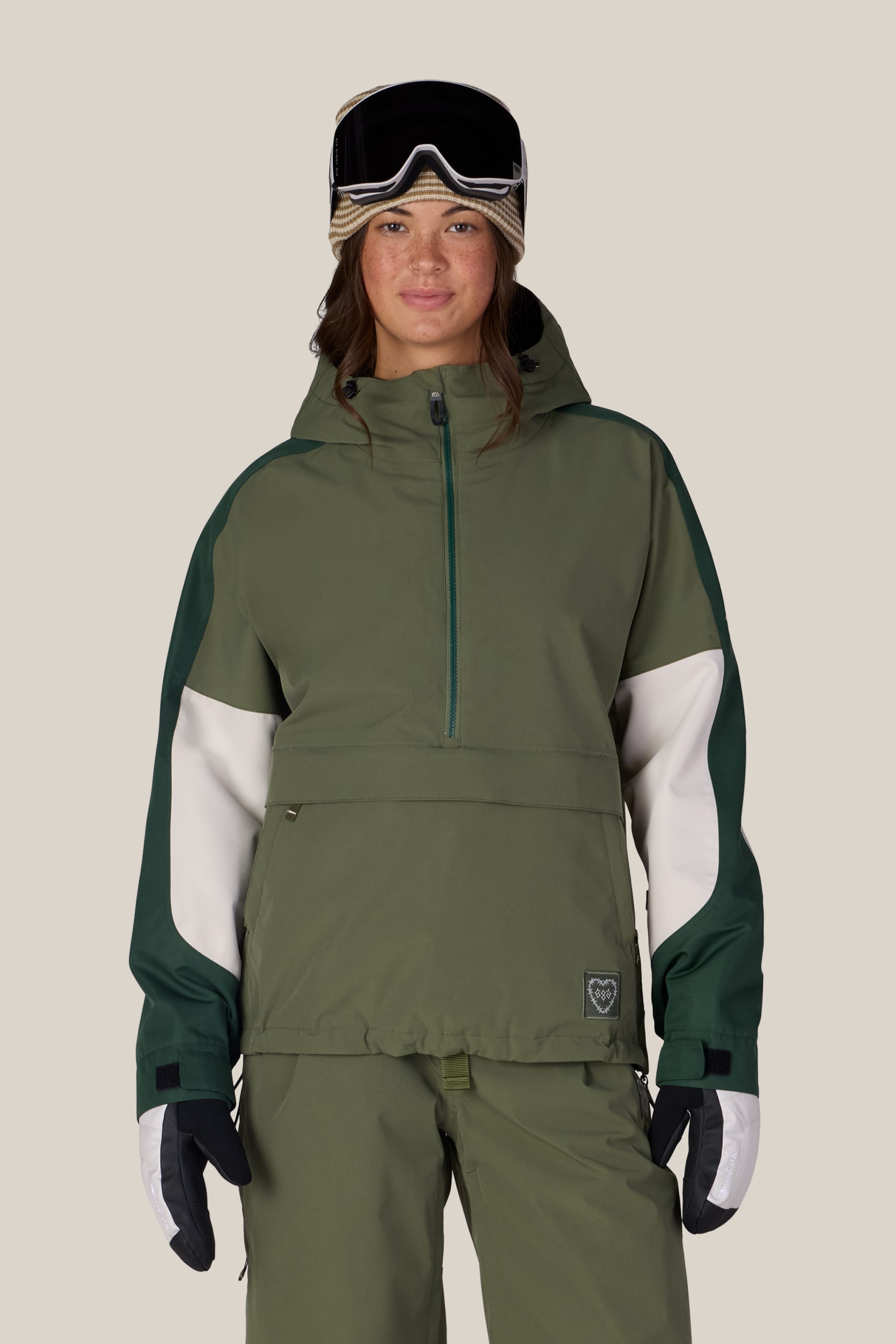 A woman wearing the 686 Women’s Outline™ Shell Anorak by 686, paired with matching green pants, black and white gloves, and ski goggles on her head stands facing forward against a plain background.
