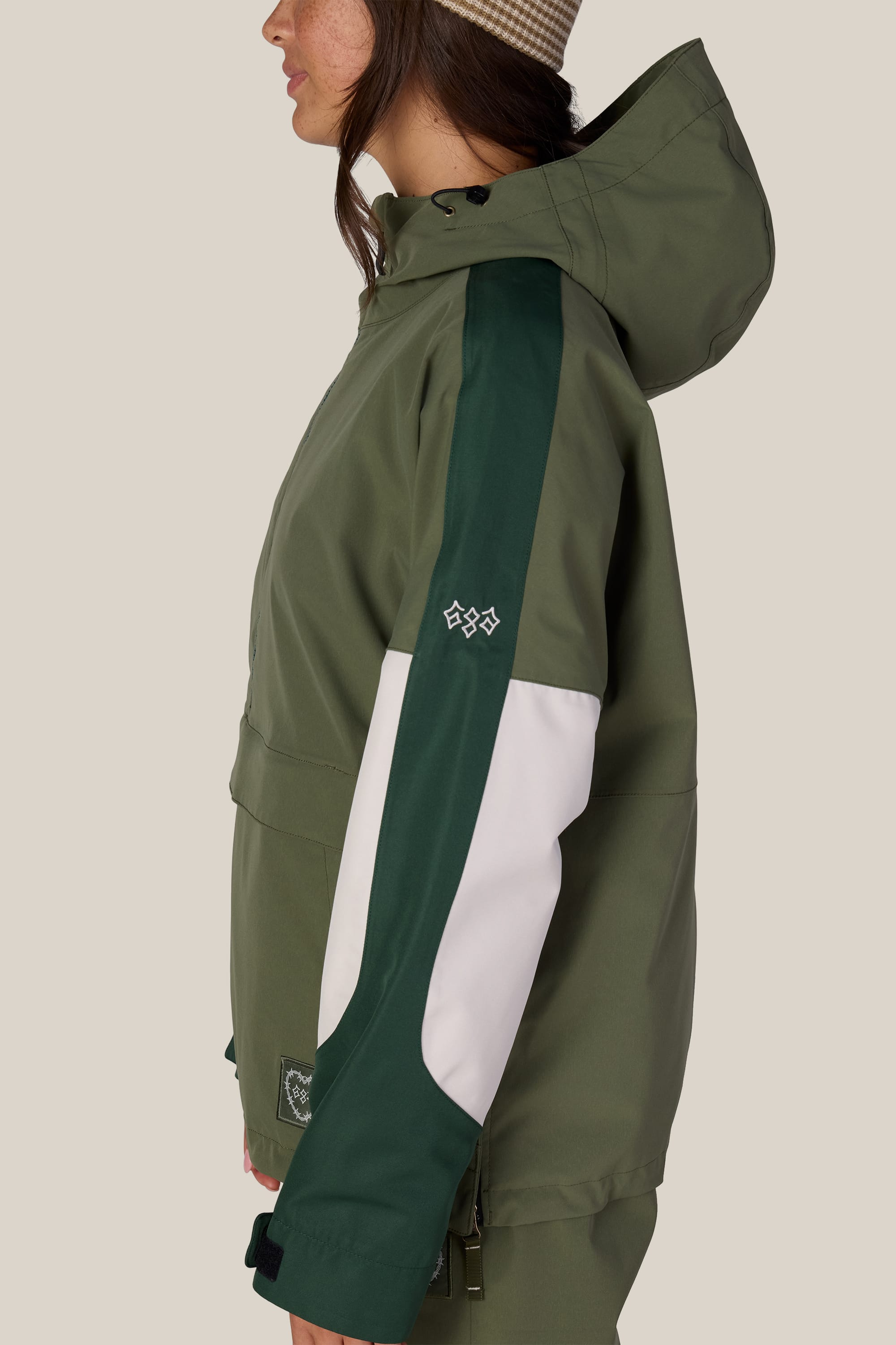 A woman is shown from the side against a plain background wearing the 686 Women’s Outline™ Shell Anorak in green, featuring a dark green and white stripe on the sleeve, paired with a beige knit hat.